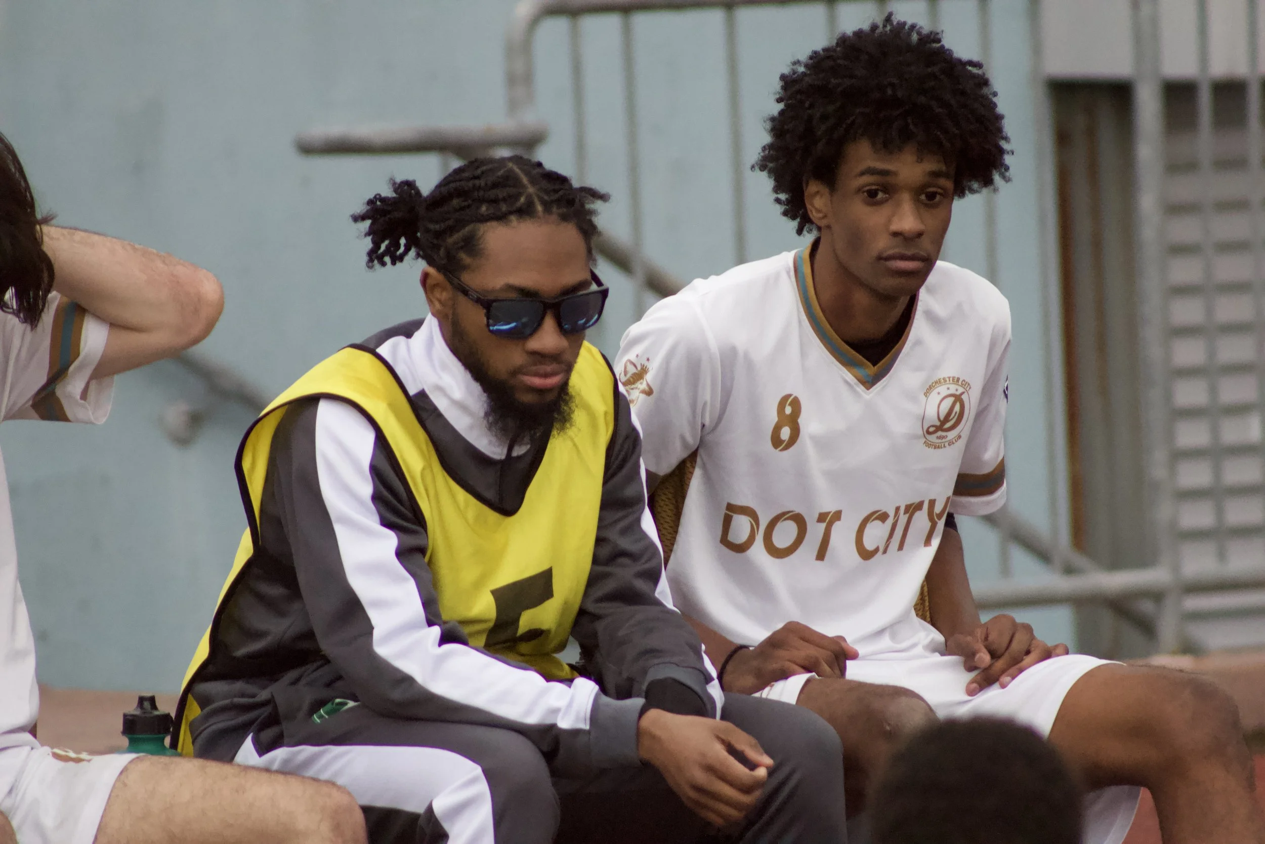 Two young men sitting on a bench, one wearing sunglasses and a yellow training vest, the other in a white sports jersey with the number 8, at a sports facility.