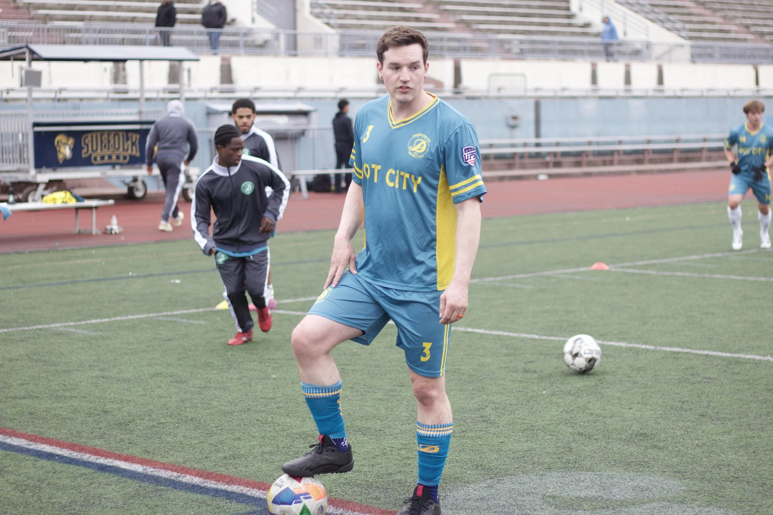 A young man in a blue yellow sports jersey, shorts, and matching socks standing on a soccer field with a soccer ball at his foot. In the background, other people are on the field and in the bleachers.