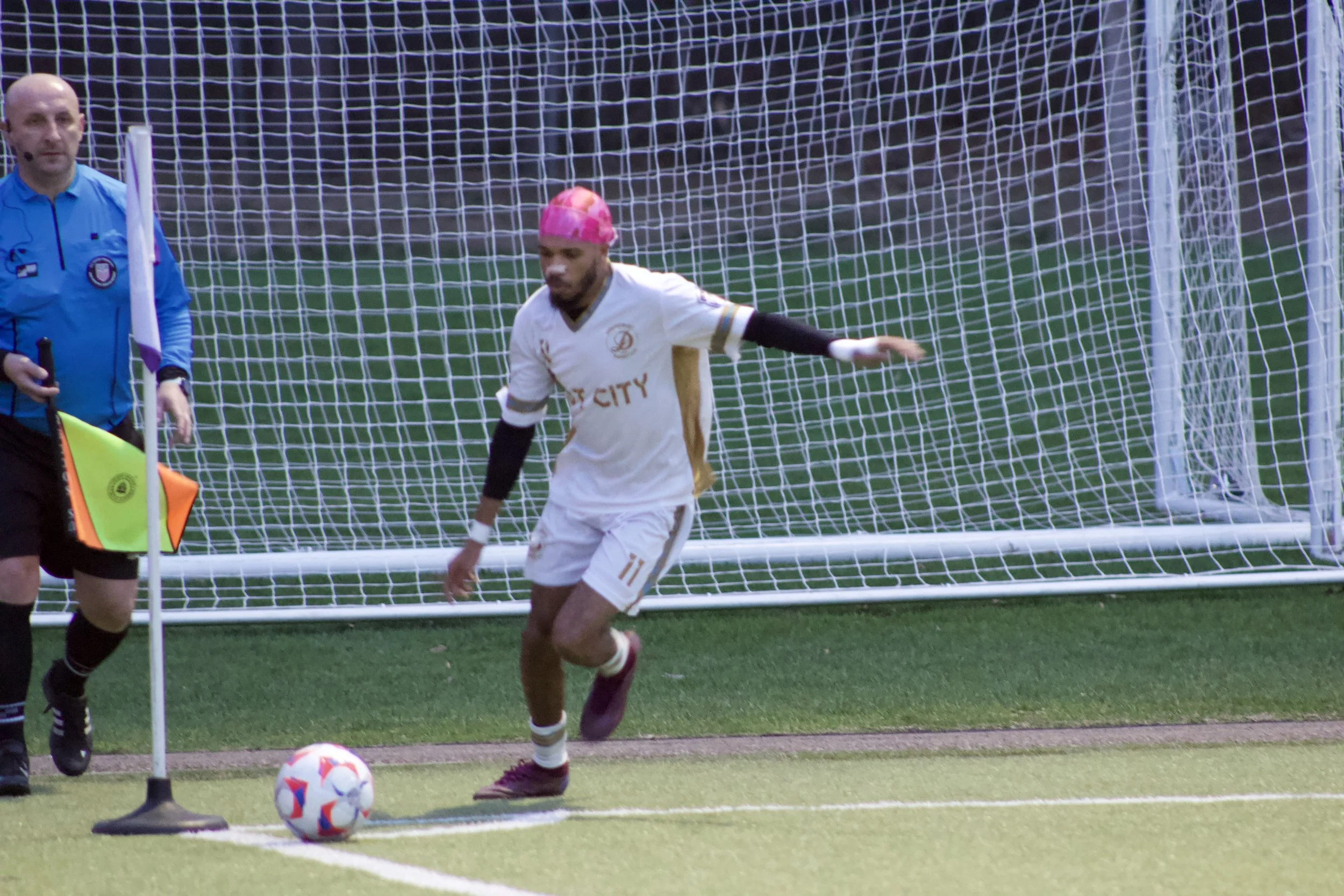 A soccer player wearing a white jersey and pink cap kicks a soccer ball near the corner flag as an official stands nearby on the field.