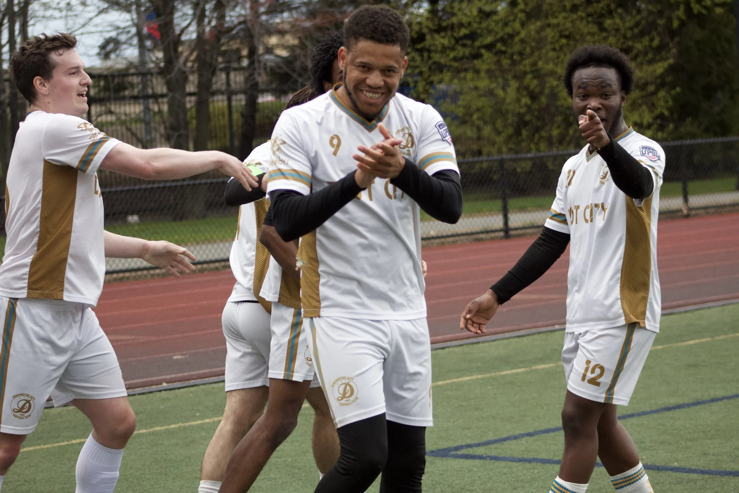 A group of soccer players in white and gold uniforms on a field, smiling and celebrating, with a chain-link fence and trees in the background.