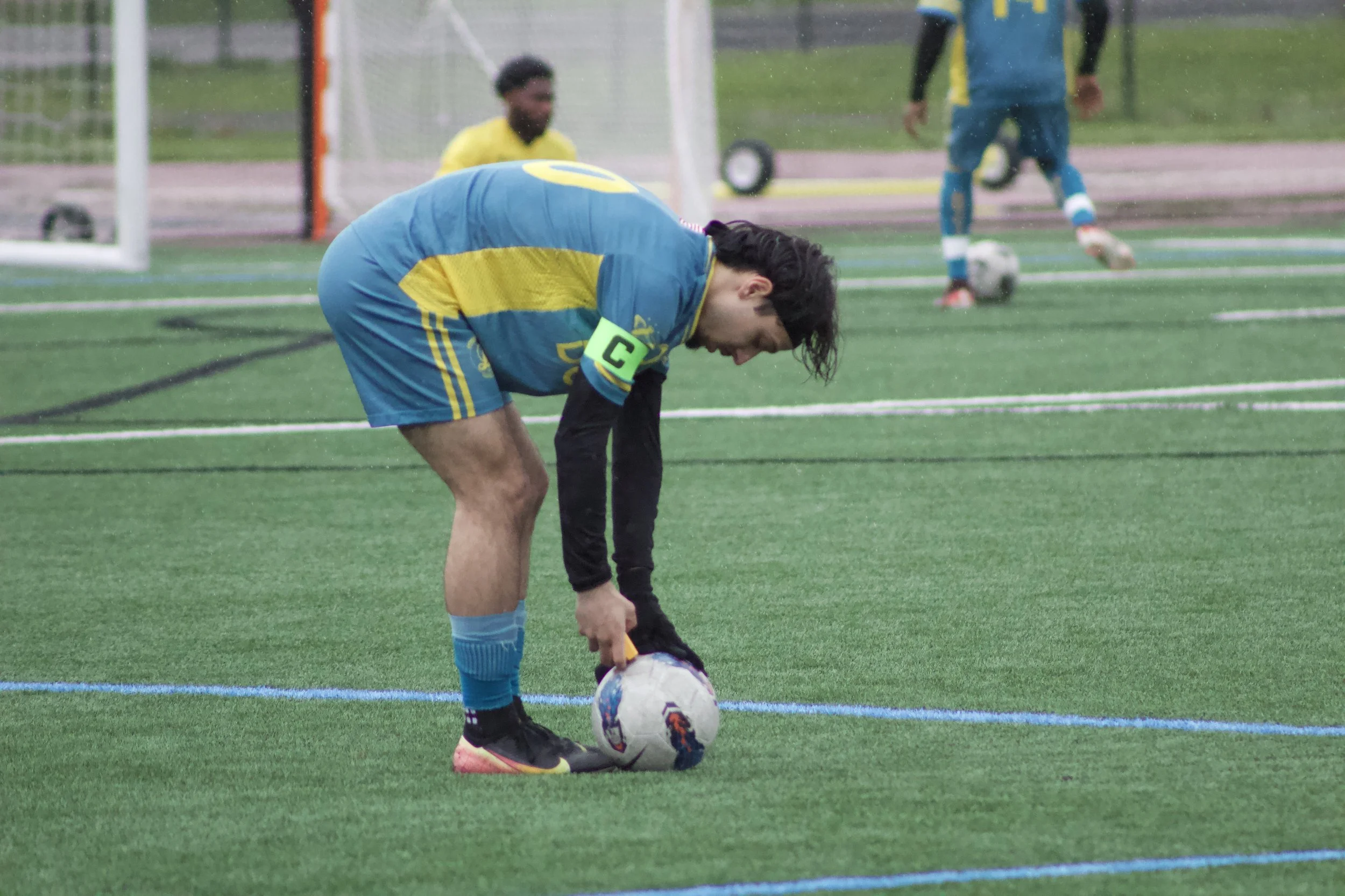 A soccer player in a blue and yellow uniform bends down to adjust his soccer cleats on a soccer field during a game or practice, with a soccer ball nearby, and a goalkeeper and another player in the background.