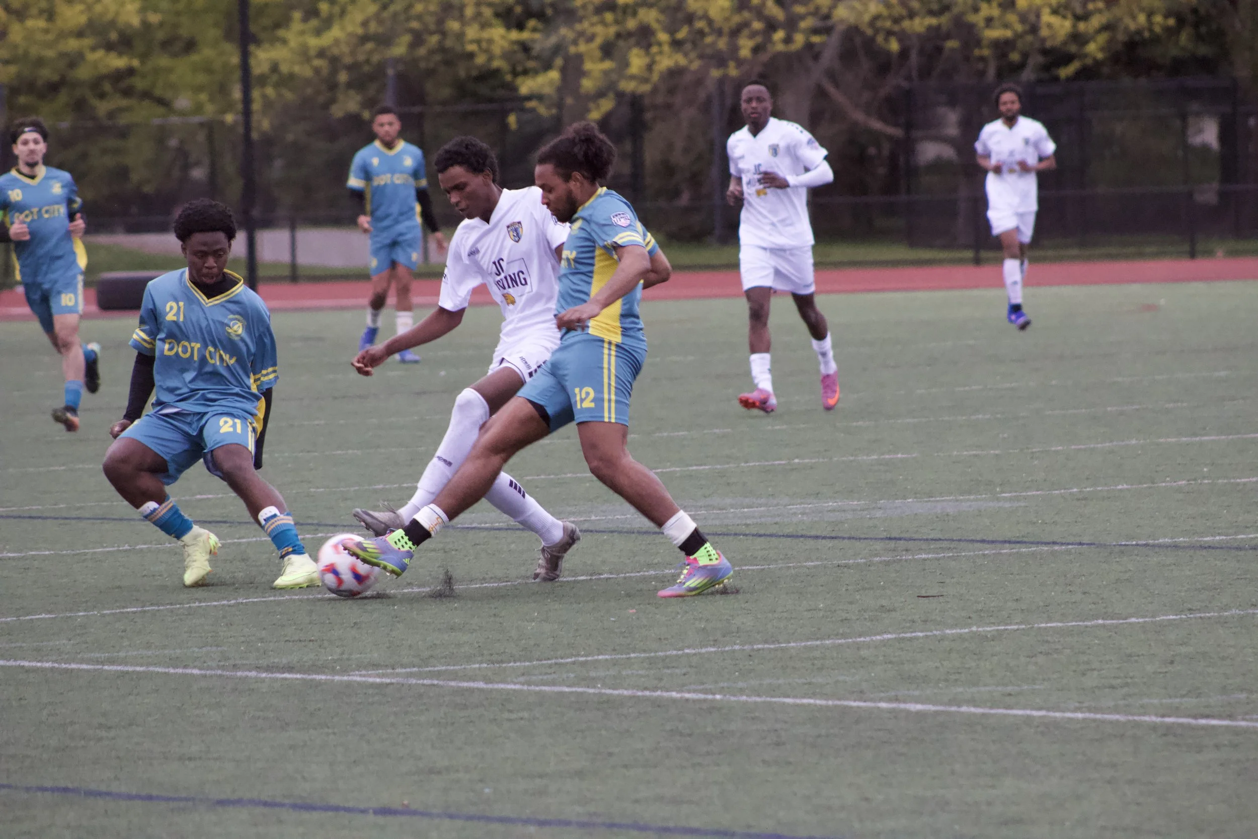 A group of soccer players, wearing blue and white jerseys, are playing a match on an outdoor field. Two players in the foreground are battling for the ball, while others watch or move in the background. Trees surround the field.
