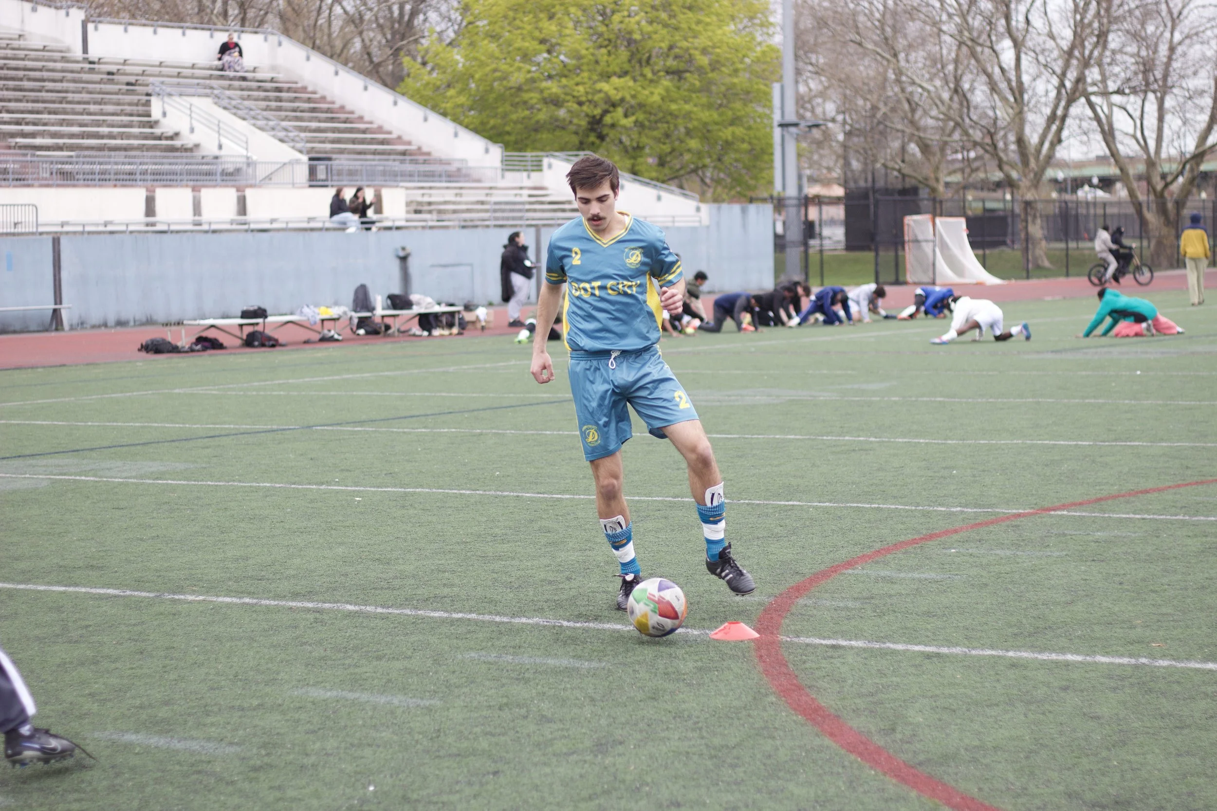 A young man dressed in a blue soccer uniform and black shoes is standing on a soccer field, preparing to kick a soccer ball. In the background, people are sitting, walking, and stretching on the sidelines, with some volunteering or coaching. There ar