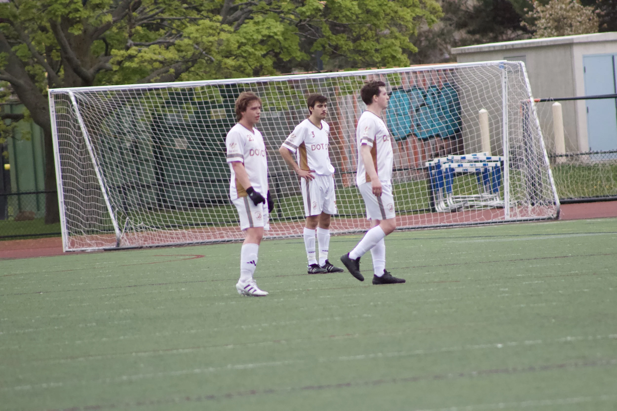 Three young male soccer players in white uniforms standing on a soccer field in front of a goal, with trees and a small building in the background.