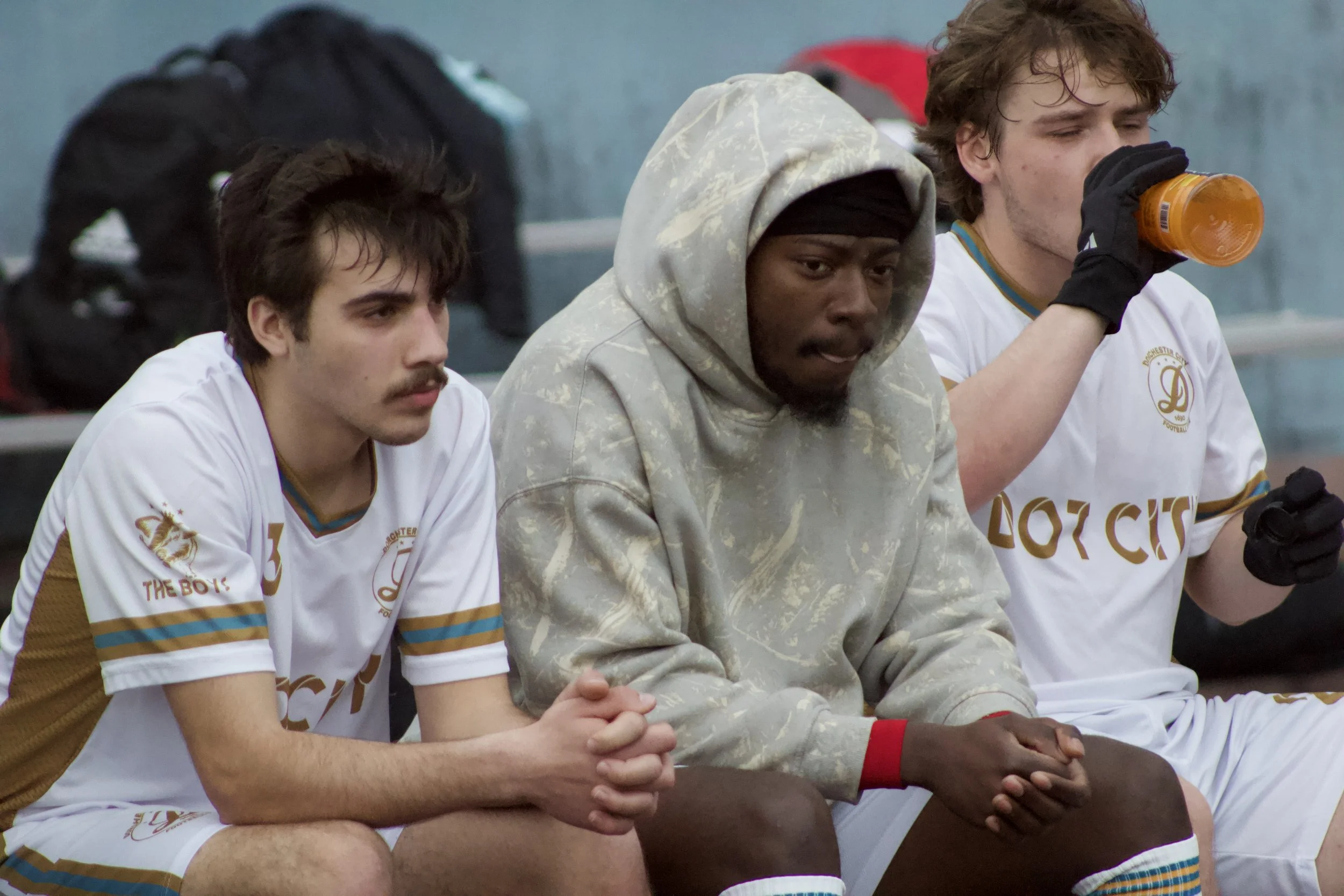 Three young men sitting on a bench, wearing sports jerseys, with one drinking from a bottle. They appear to be tired or contemplative, possibly during a sports event.