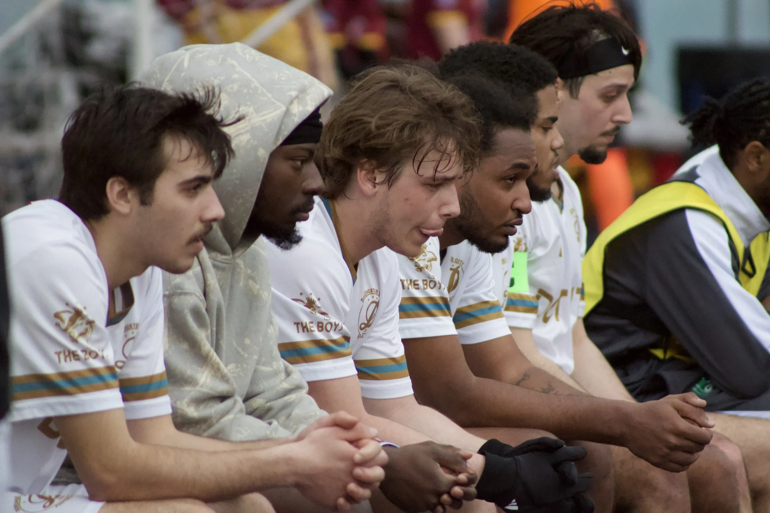 Six young men sitting side by side, with heads bowed and hands clasped, appear to be in a moment of silence or prayer, wearing sports jerseys and casual attire.