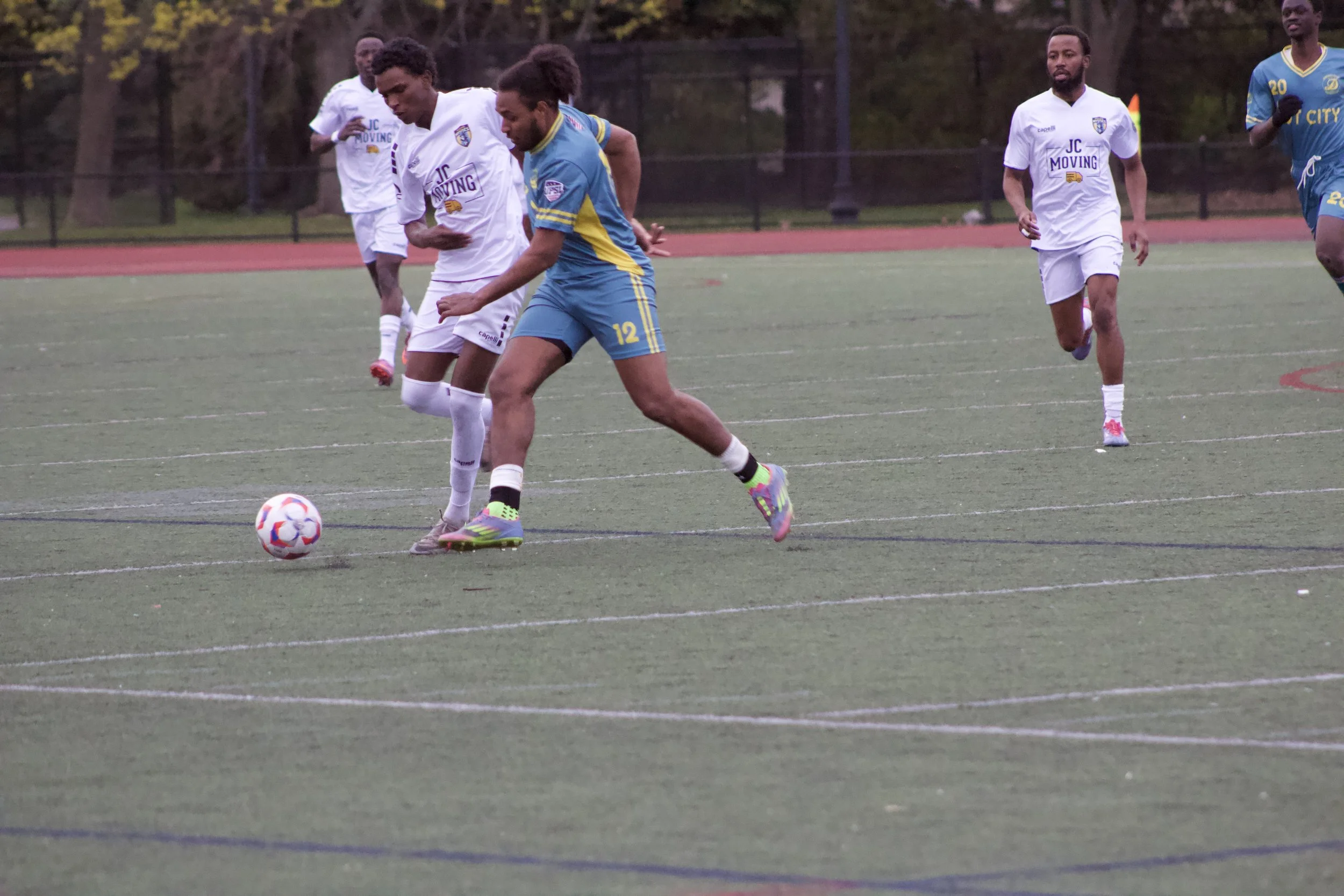 A group of young men playing soccer on a green field with trees in the background. One player is about to kick the ball, wearing a blue and yellow jersey, while others in white jerseys are running nearby.