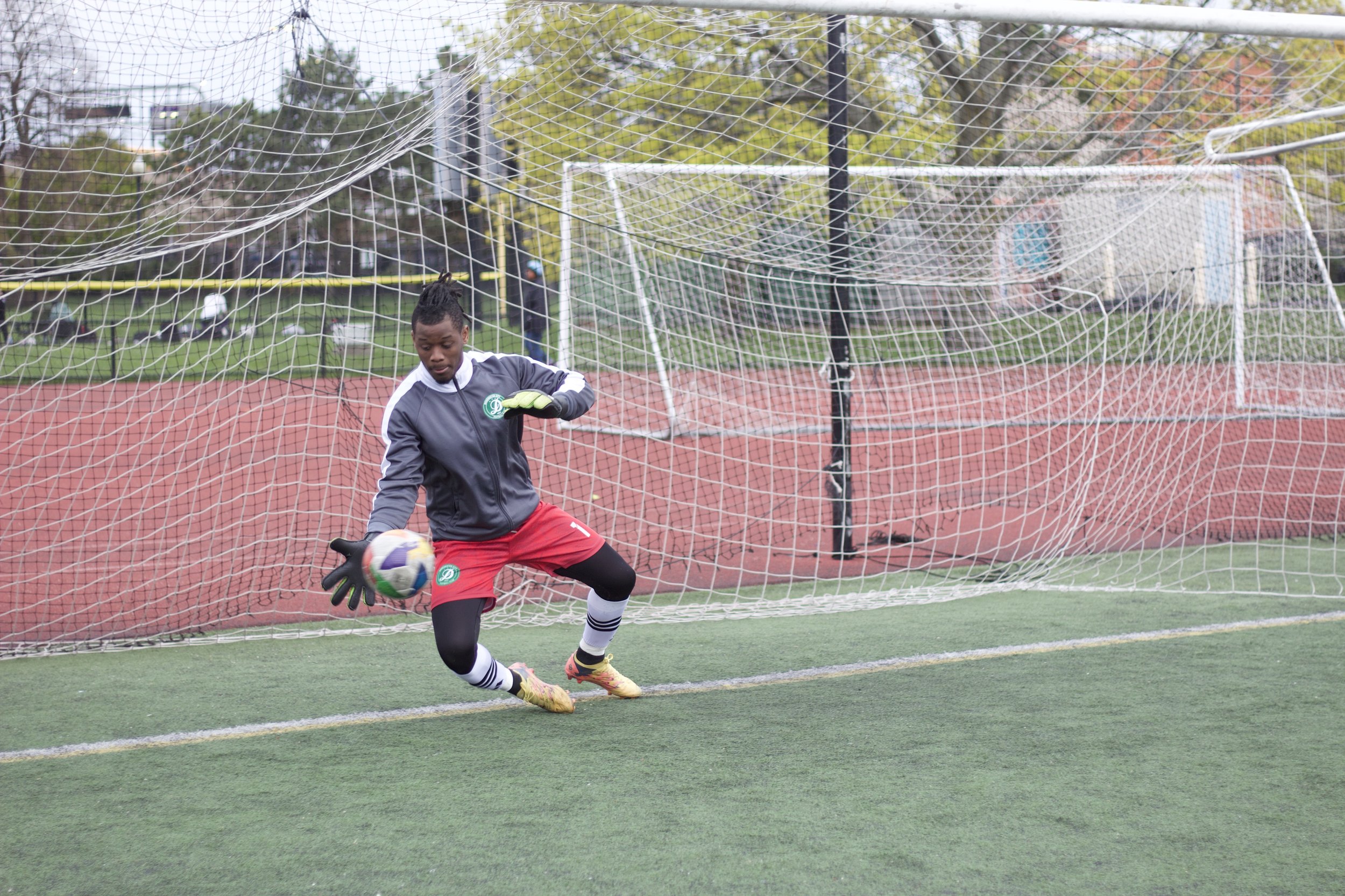 A soccer goalie in a black and red uniform catching a soccer ball on a field with a net and trees in the background.