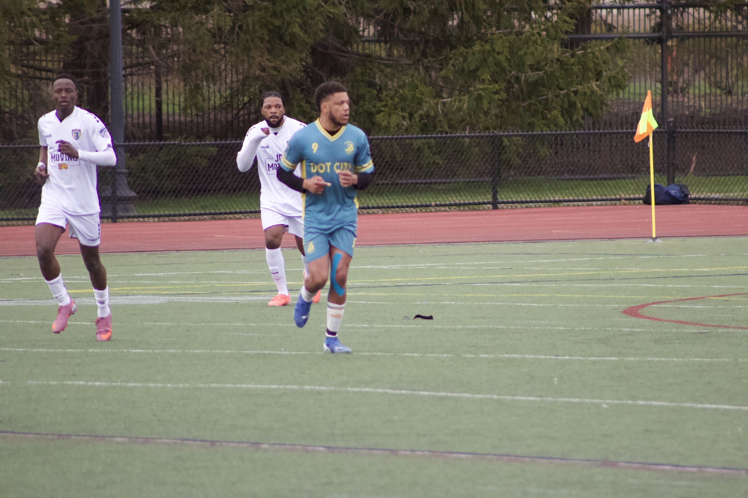 Three soccer players on a field, with two wearing white jerseys and one in a blue jersey, near a corner flagpost.