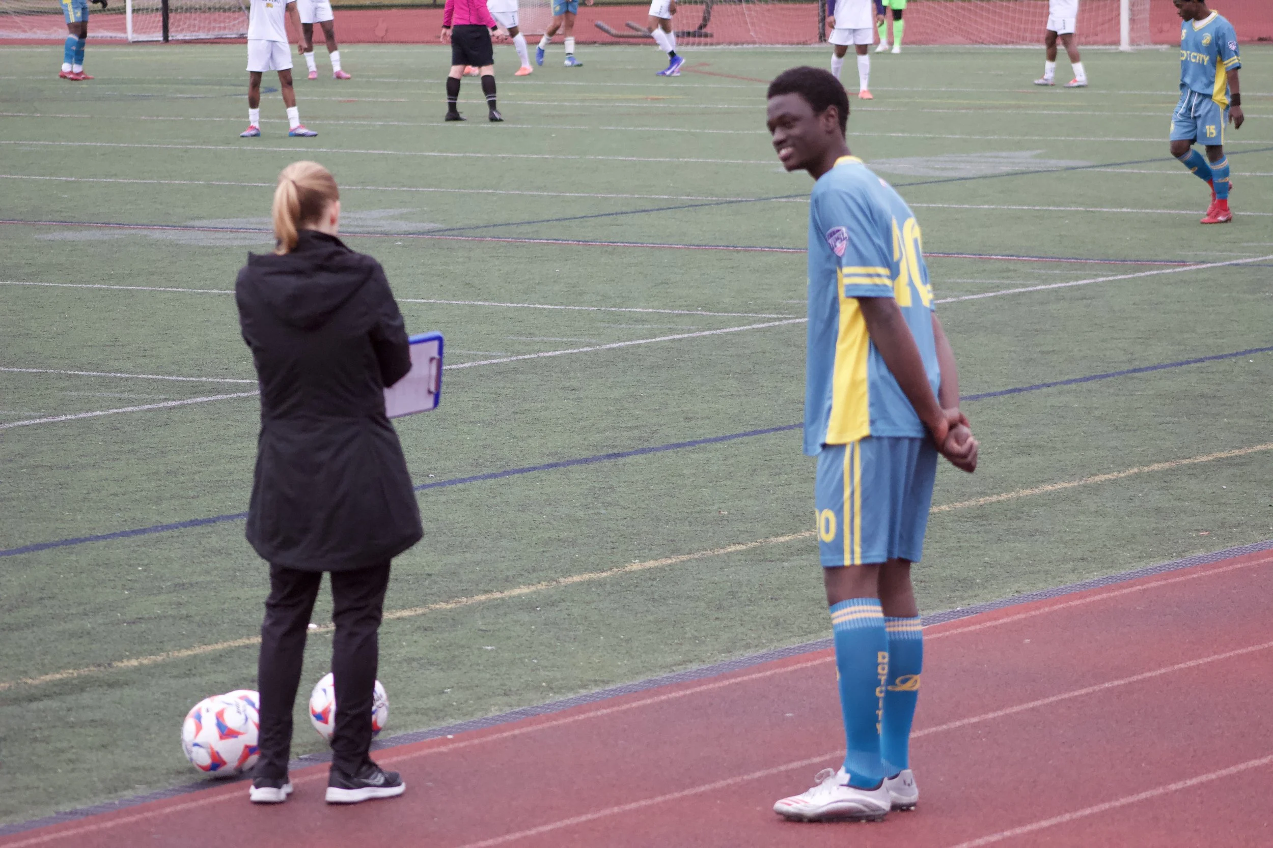 A soccer player in a blue uniform standing on the sideline of a soccer field, observing a woman with a clipboard, with other players and a referee on the field in the background.