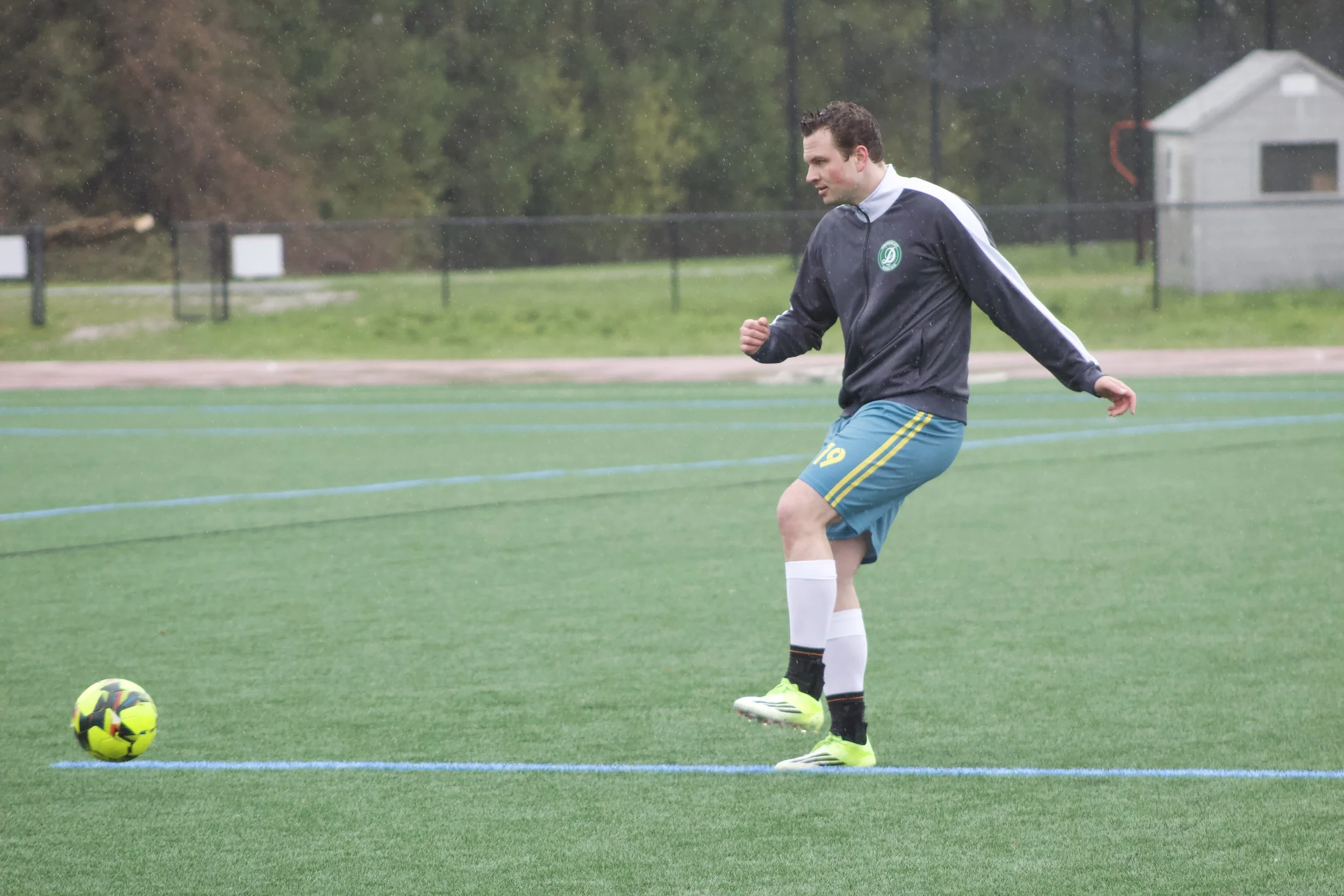 A man in a soccer uniform is on a grassy soccer field, about to kick a soccer ball in the rain.