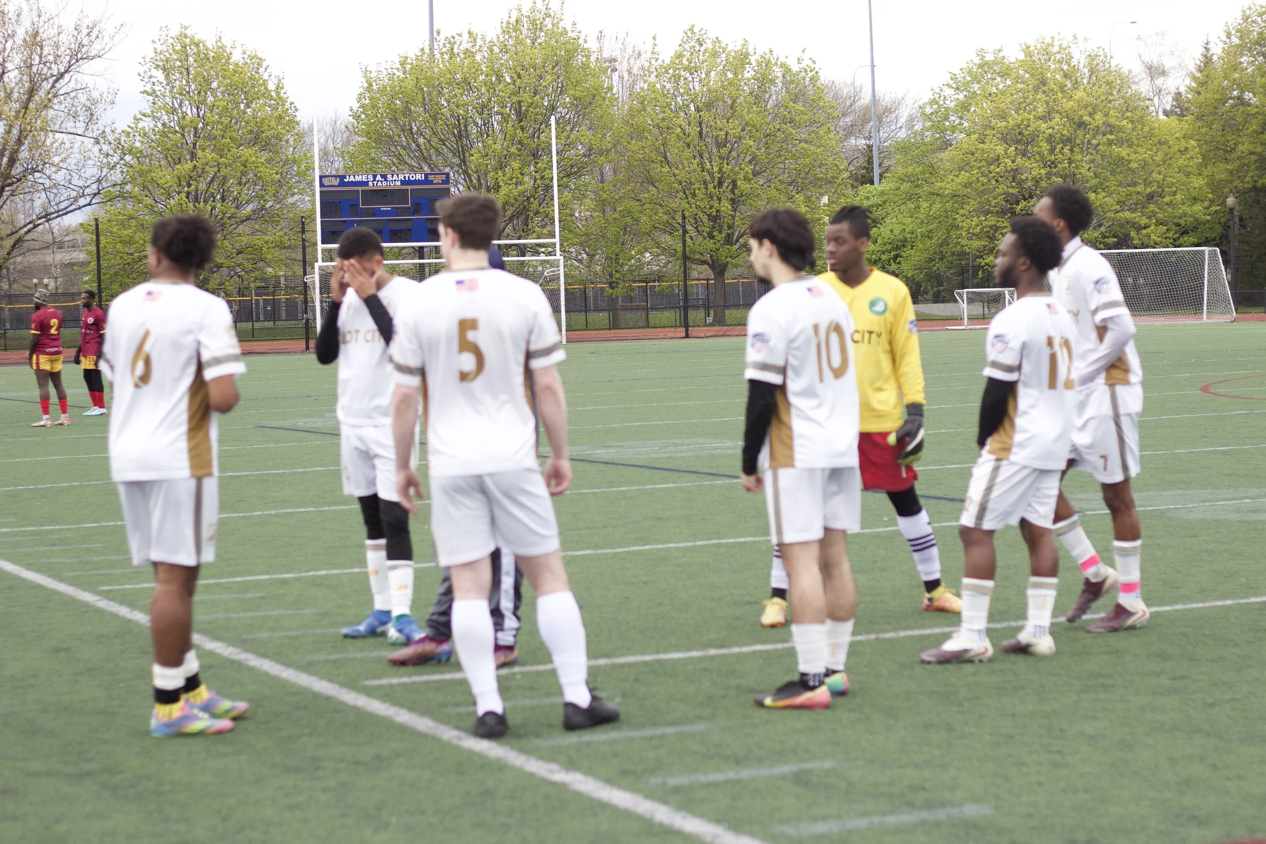 A group of soccer players, wearing white and yellow uniforms, standing on a soccer field with green grass, trees, and a scoreboard in the background.