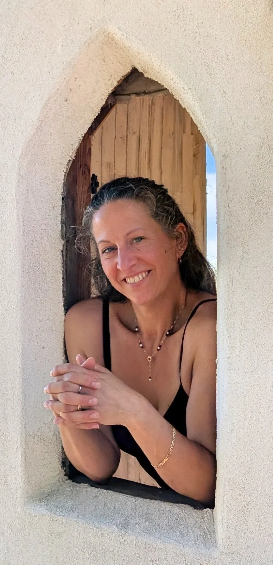 A woman with dark, wavy hair smiling and leaning through a small, arched opening in a wall. She is wearing a black spaghetti strap top, jewelry, and has her hands clasped together.