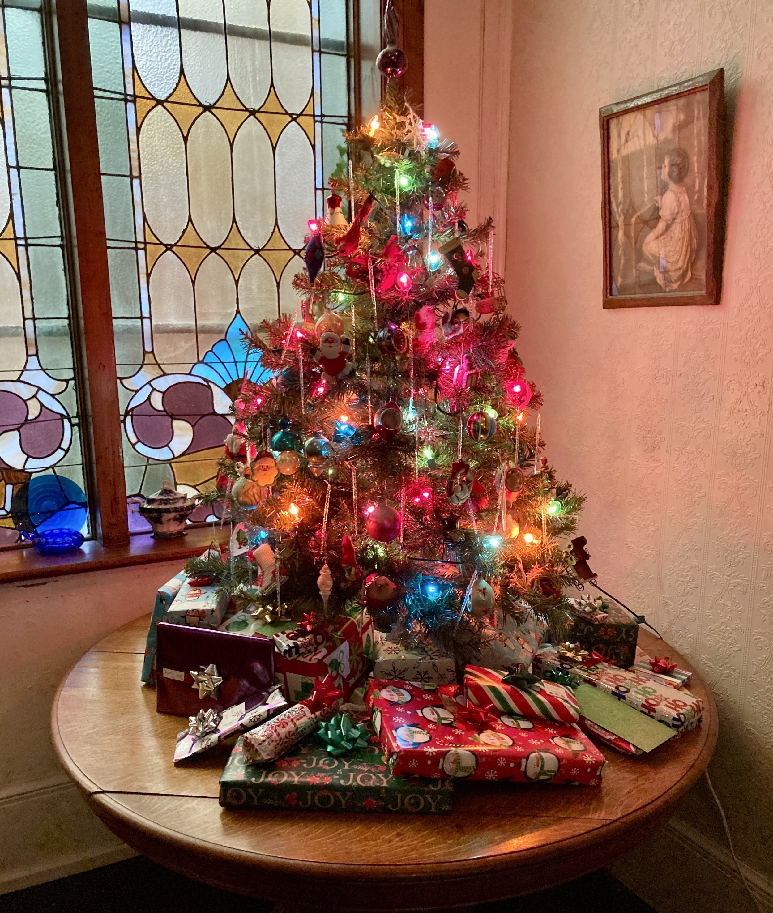 A decorated Christmas tree with colorful lights, ornaments, and toys, surrounded by wrapped presents on a wooden table in a room with stained glass windows and framed artwork on the walls.