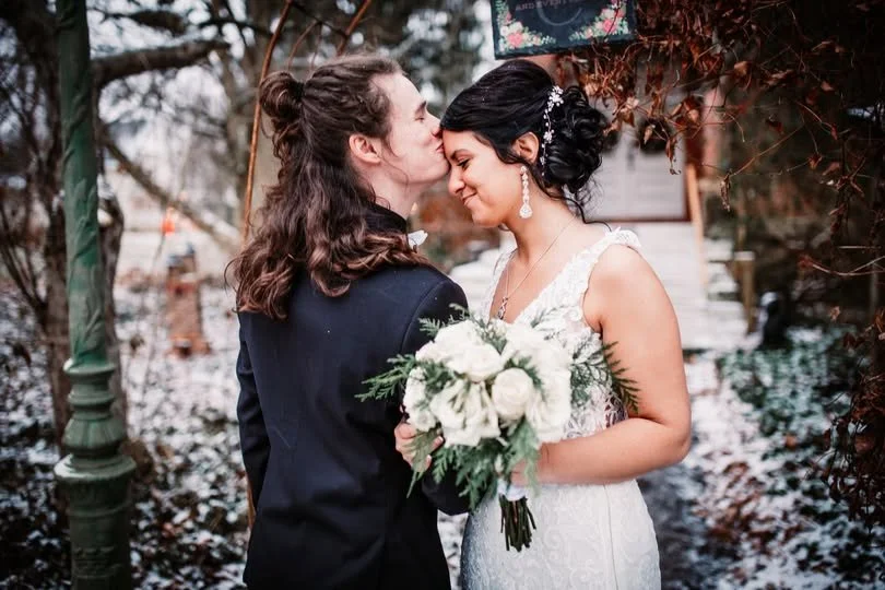 A bride and groom sharing a moment outdoors, with the bride holding a bouquet of white flowers, and surrounded by winter foliage.