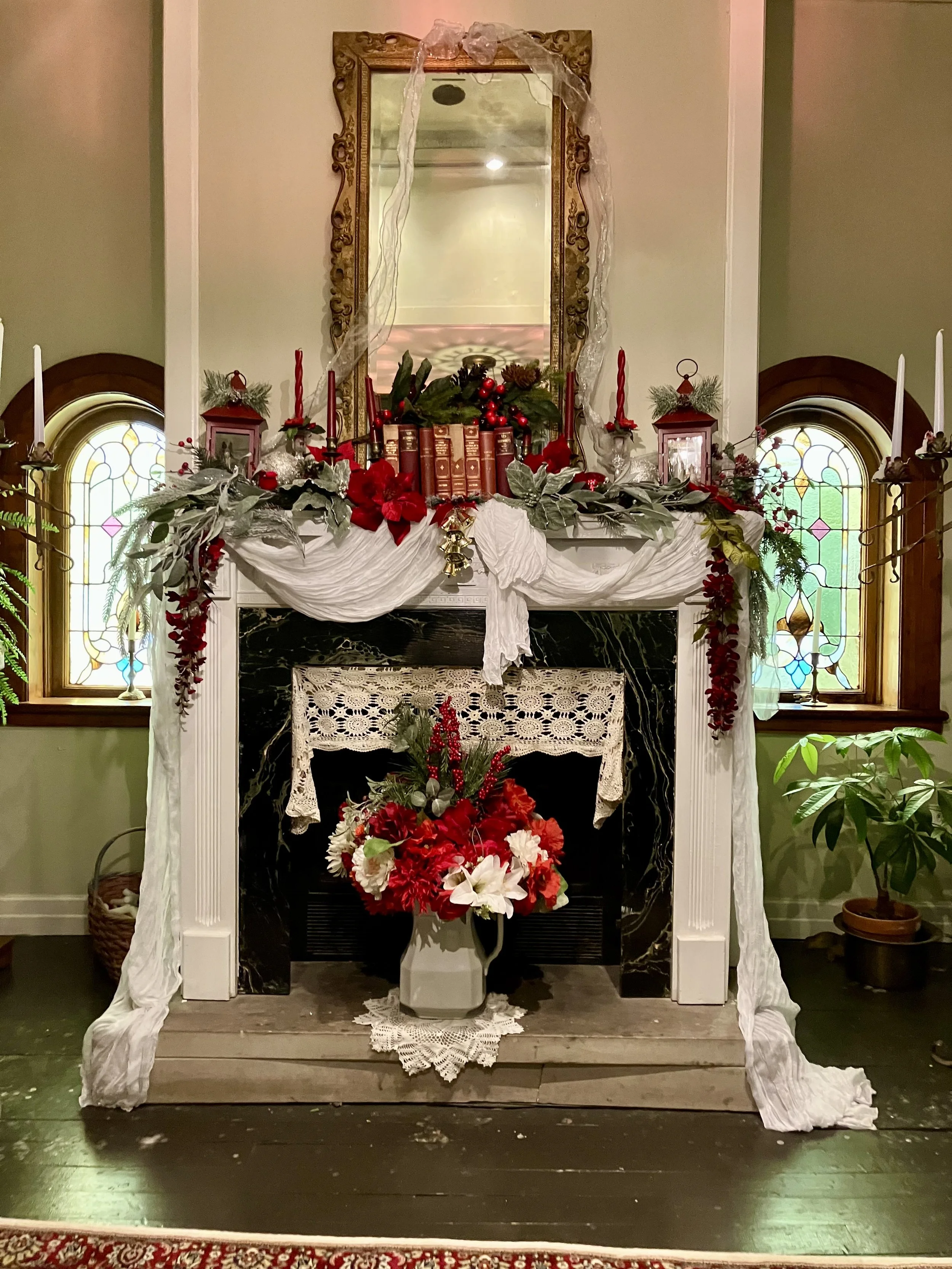 A decorated fireplace with festive Christmas decor, including a mirror with a gold ornate frame, red candles, lanterns, greenery, red and white flowers, and holiday ornaments. Two stained glass windows on either side of the fireplace.