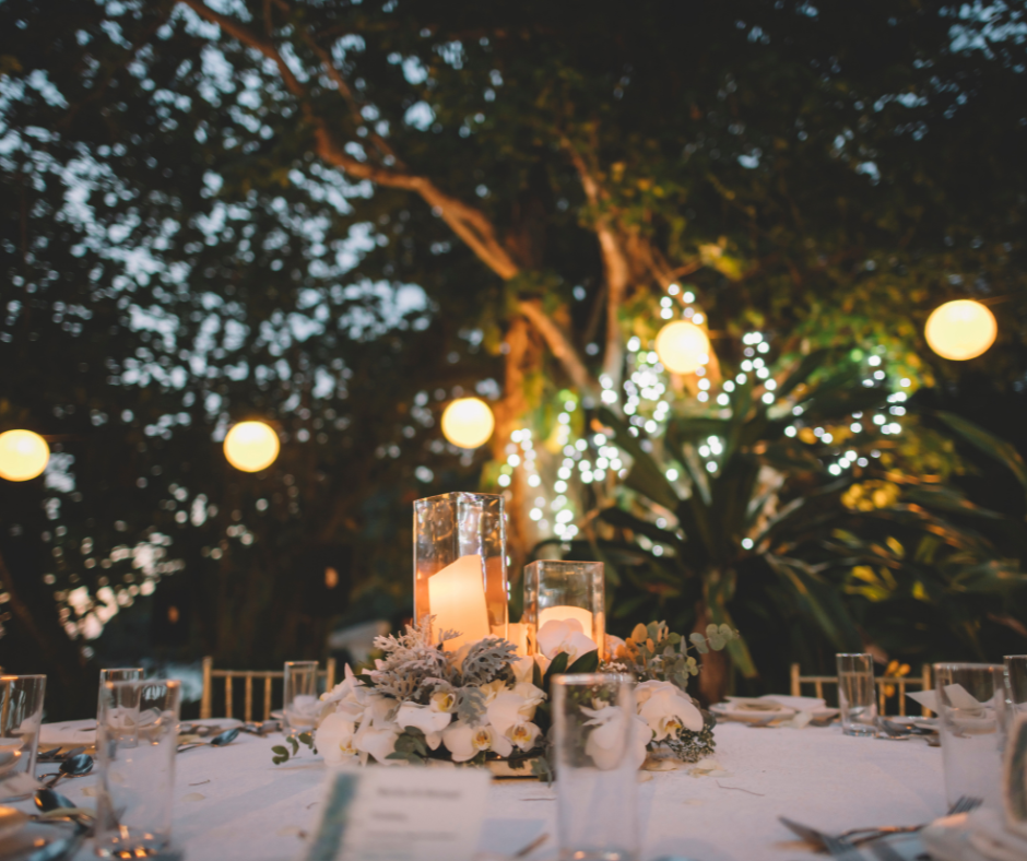 Outdoor wedding reception table with candles and floral centerpiece, surrounded by trees with string lights.