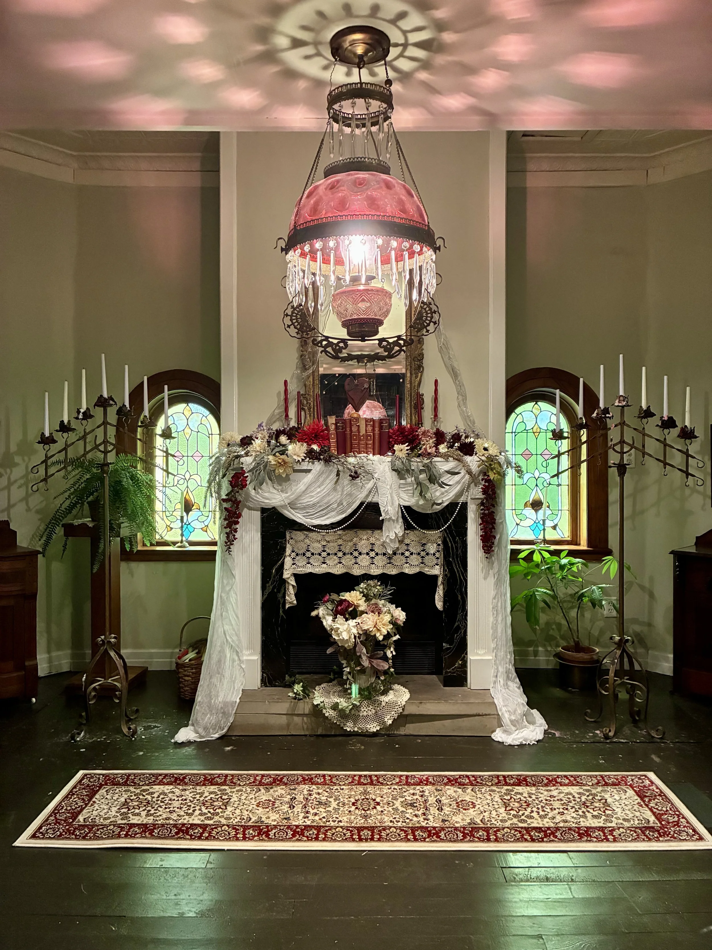 An antique chandelier casting pink and white light over a decorated fireplace mantel with flowers, candles, and a mirror, flanked by stained glass windows and green plants.