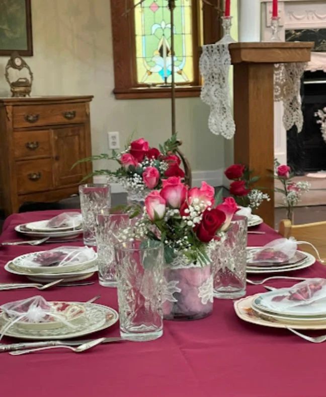 Elegant dining table setup with red tablecloth, crystal glassware, floral centerpieces with red roses, and decorative plates in a cozy room with stained glass window.