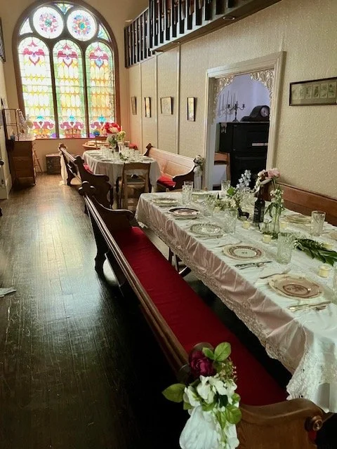 A long dining table set for a meal with plates, glasses, and flowers, in a room with stained glass windows and vintage furniture.
