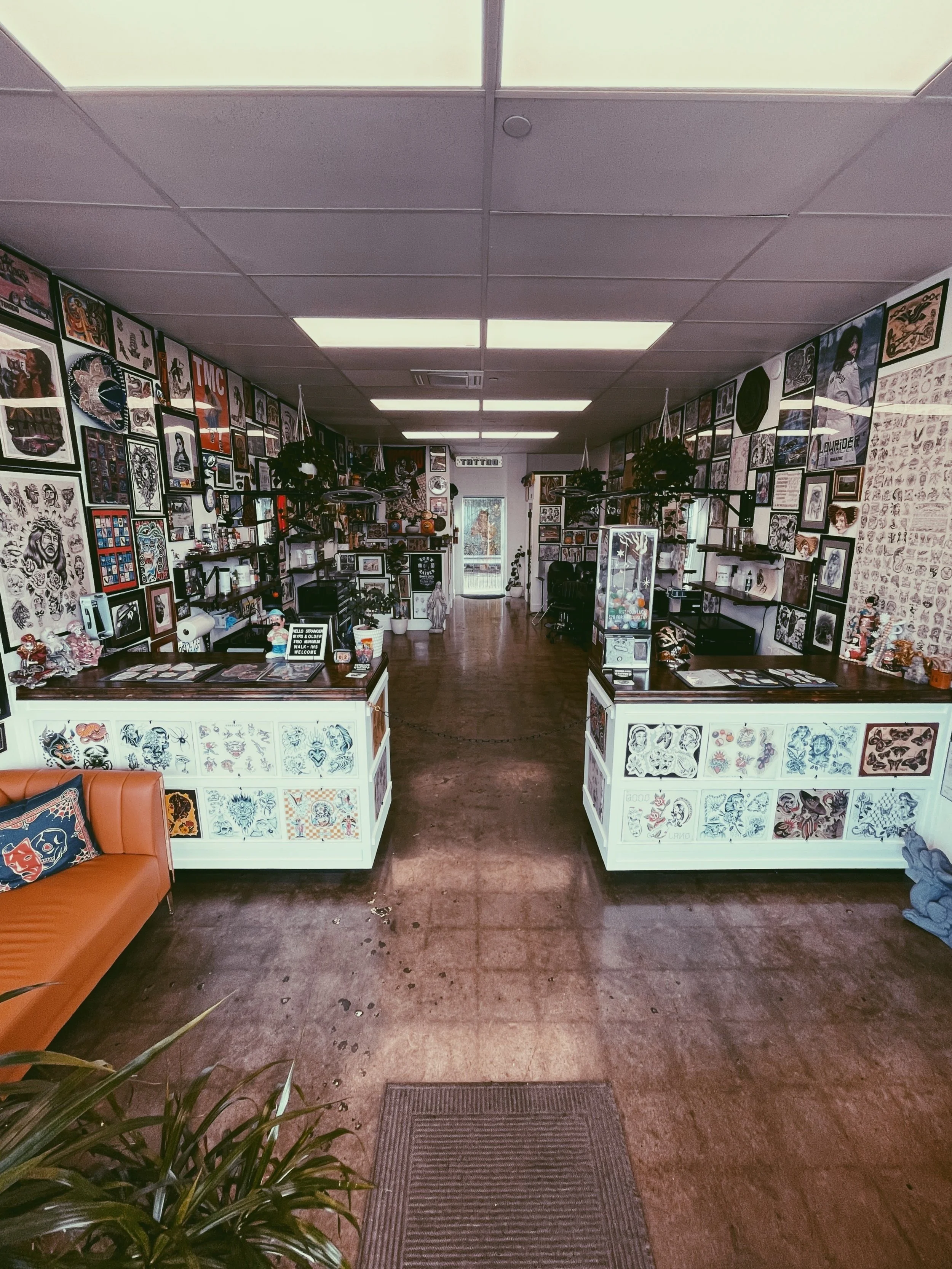View of an art studio or tattoo parlor with wall art and framed tattoo designs, a reception desk with tattoo flash, sale sign, and indoor plants, and a leather couch in the corner.