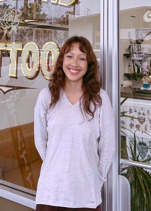 A young woman with long, curly reddish-brown hair and a big smile, standing in front of a glass window at a tattoo shop.