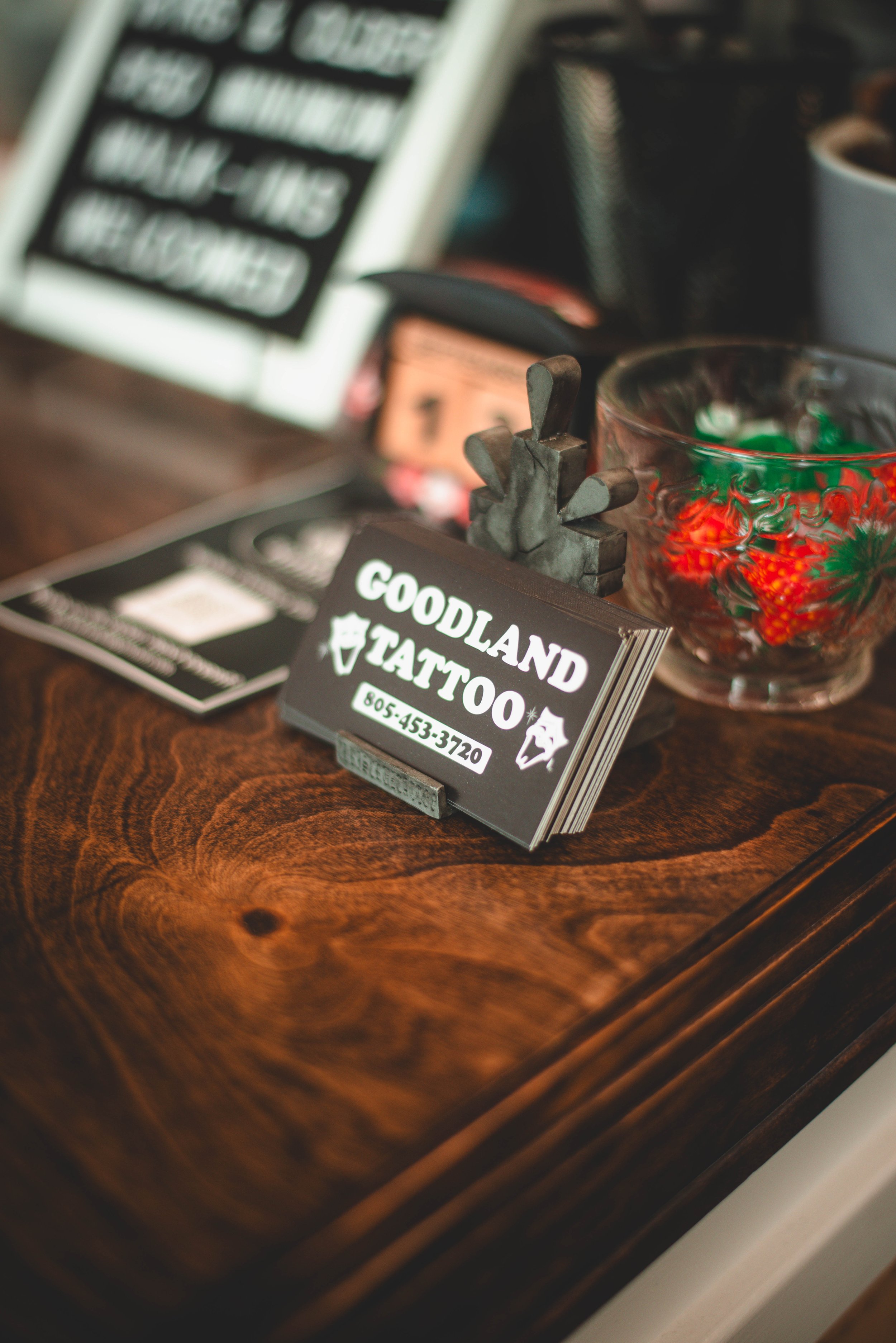 Close-up of a wooden surface with a sign, a glass bowl with candy, and a business card for goodland tattoo.