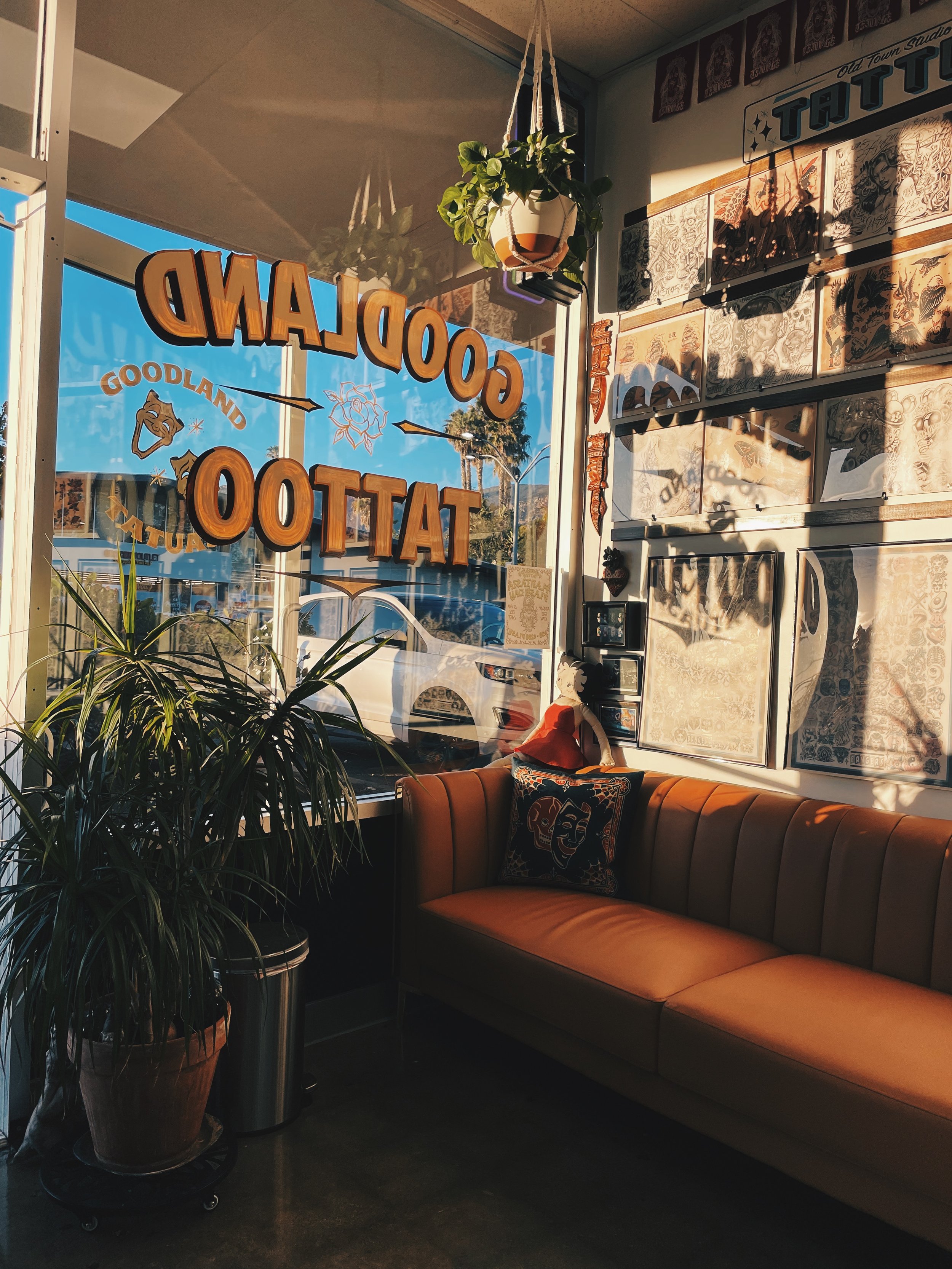 View from inside tattoo studio showing large window with 'CLOSED' sign, potted plants, and framed tattoo art on walls, with a tan leather couch and decorative pillows nearby.