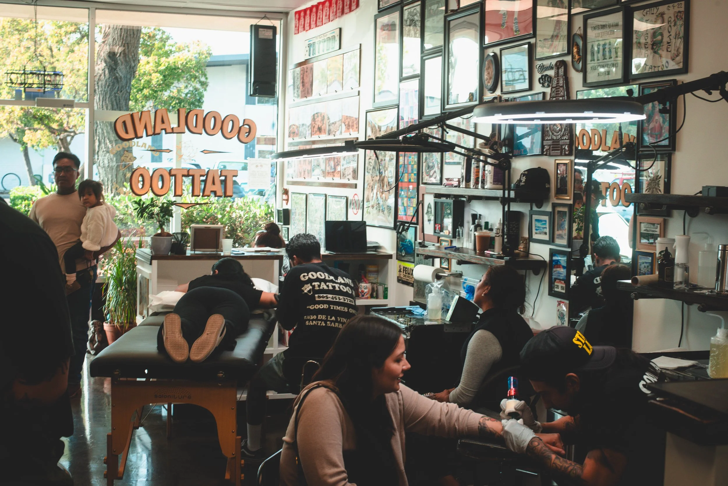 Inside a tattoo shop with framed artwork on the walls, several people seated, one person with tattoos getting work done, and a person holding a child standing near a window with sunlight and trees outside.