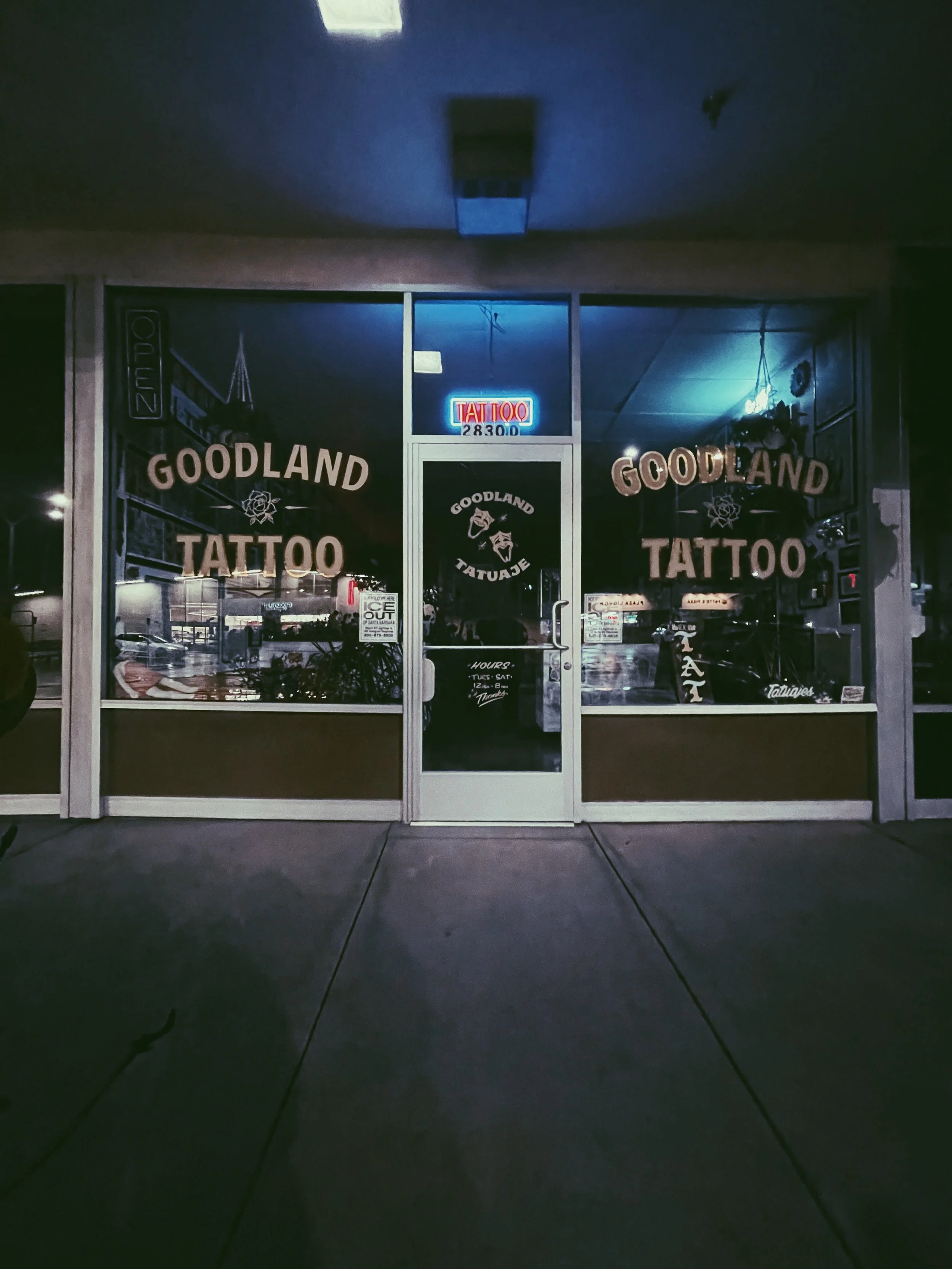 The storefront window of Goodland Tattoo is illuminated with neon signs and displays various tattoo design posters. The door is closed, and reflections of the street and parked cars are visible in the window.