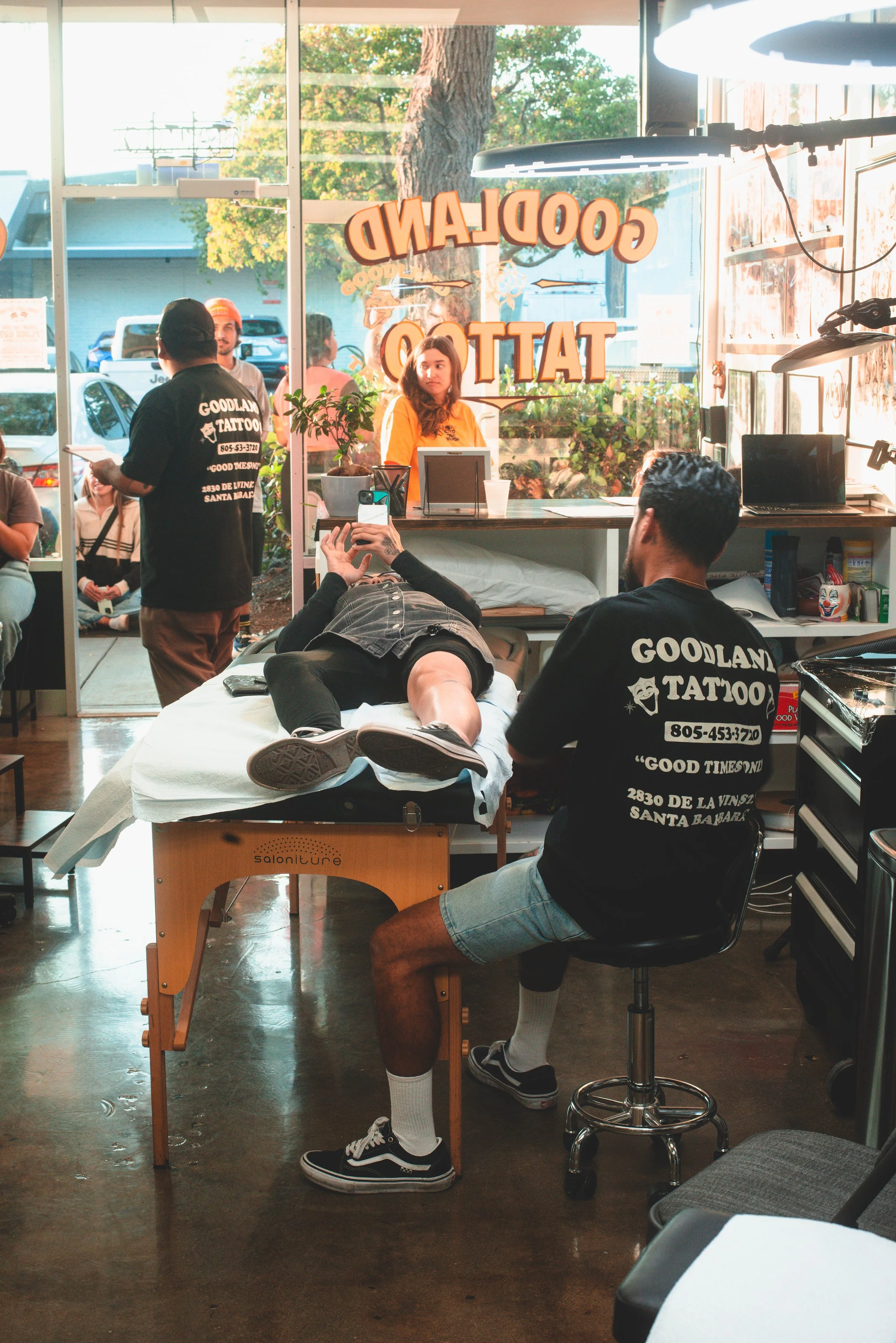 A person lying on a medical table with their shirt lifted, while another person in a black shirt with 'Goodland Tattoo' printed on the back points at their arm inside a tattoo studio. There are other people visible outside through the window, and the studio has a counter with plants and a computer.