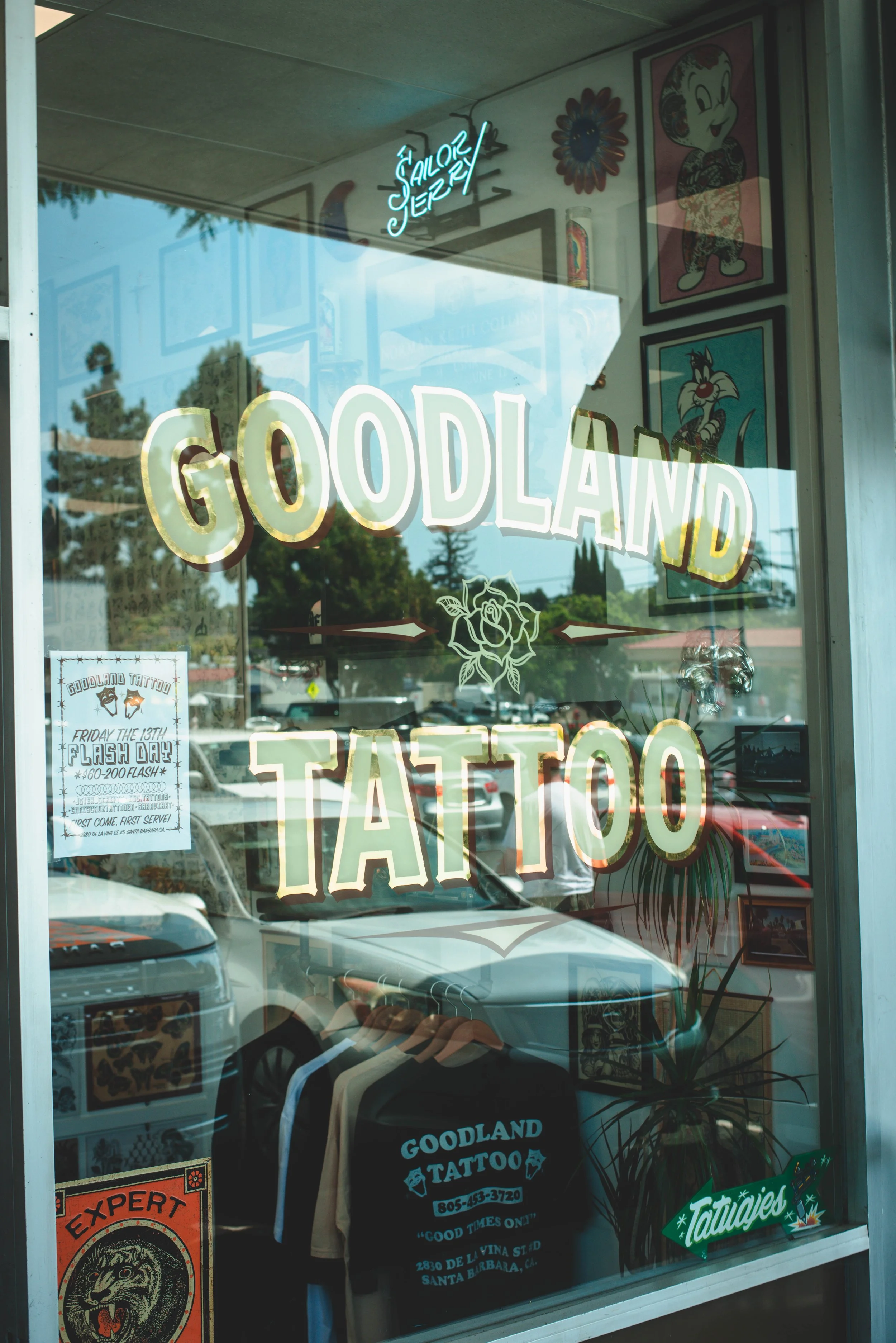 Storefront window of Goodland Tattoo shop with large white and gold lettering, tattoo-themed posters, a neon sign, and clothing on display inside.