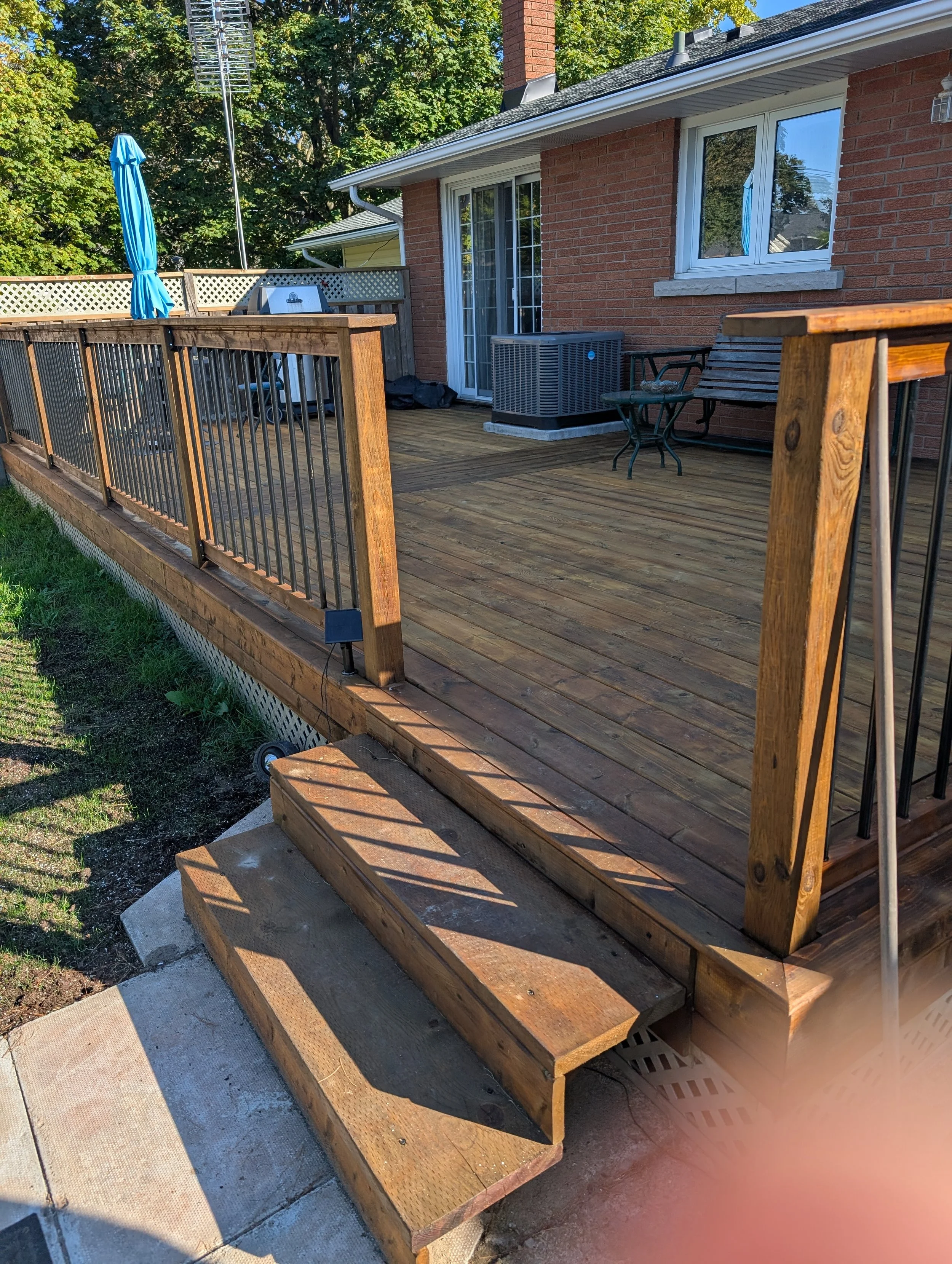 A newly built wooden deck attached to a red brick house with sliding glass door, patio furniture, and a blue umbrella on the patio, surrounded by trees.