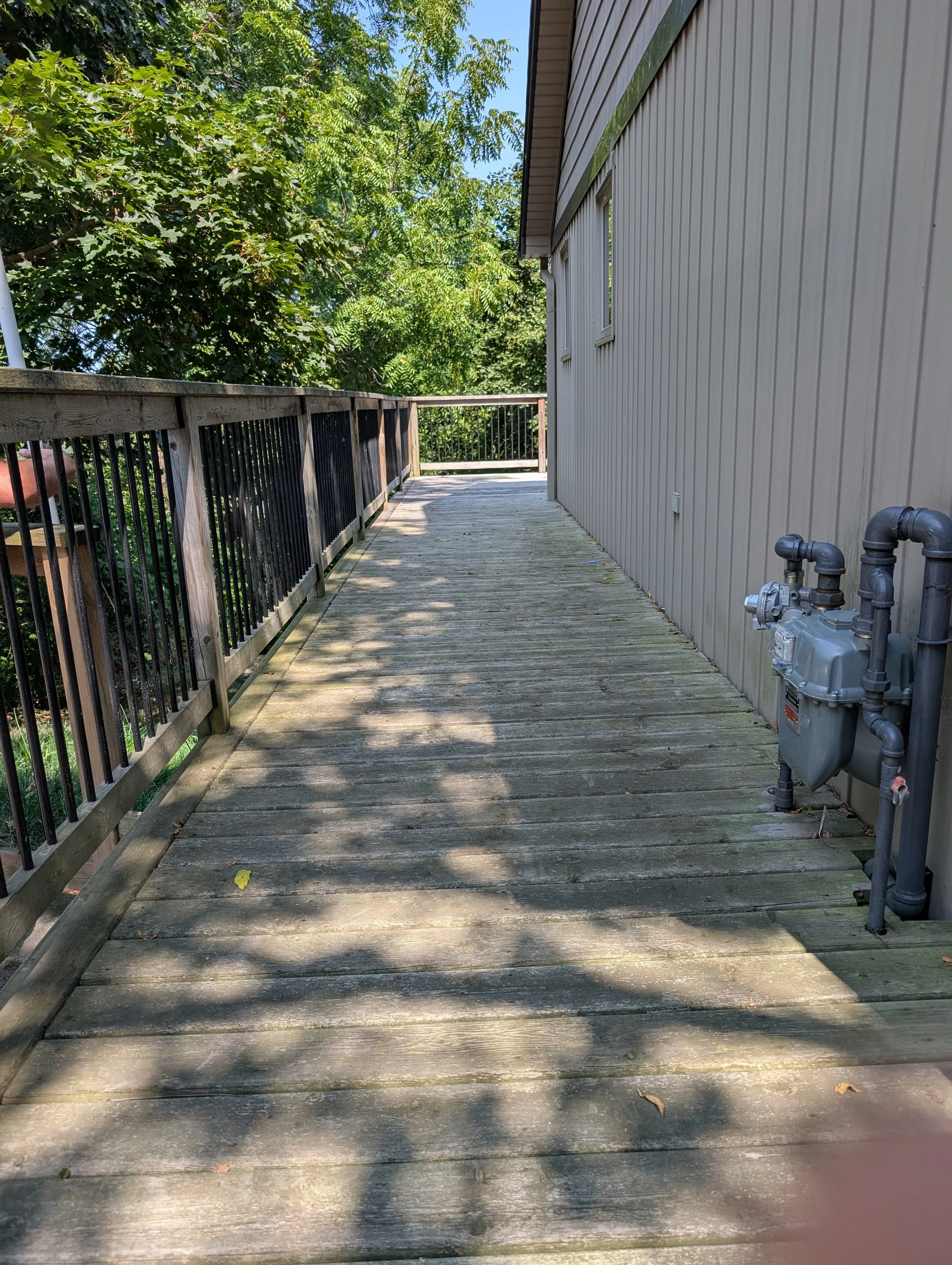 A wooden deck next to the side of a house with a utility meter, with trees and blue sky in the background, and shadows from the trees on the deck.