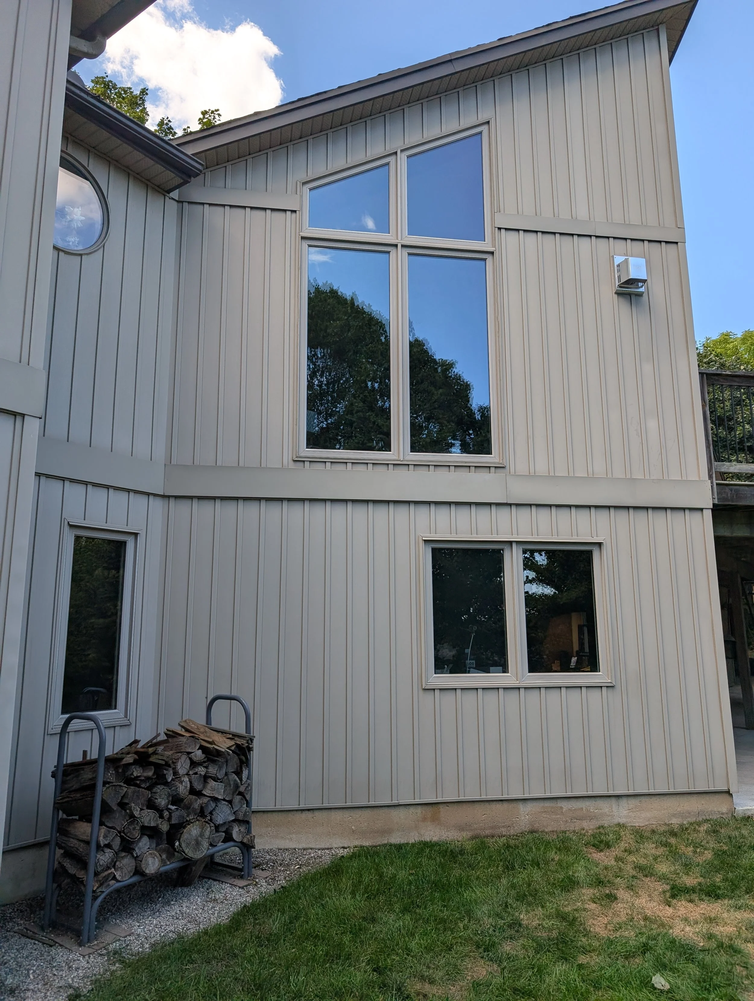 The exterior of a modern house with beige vertical siding, large windows reflecting trees and sky, and a small woodpile with logs stacked on a metal holder in the yard.