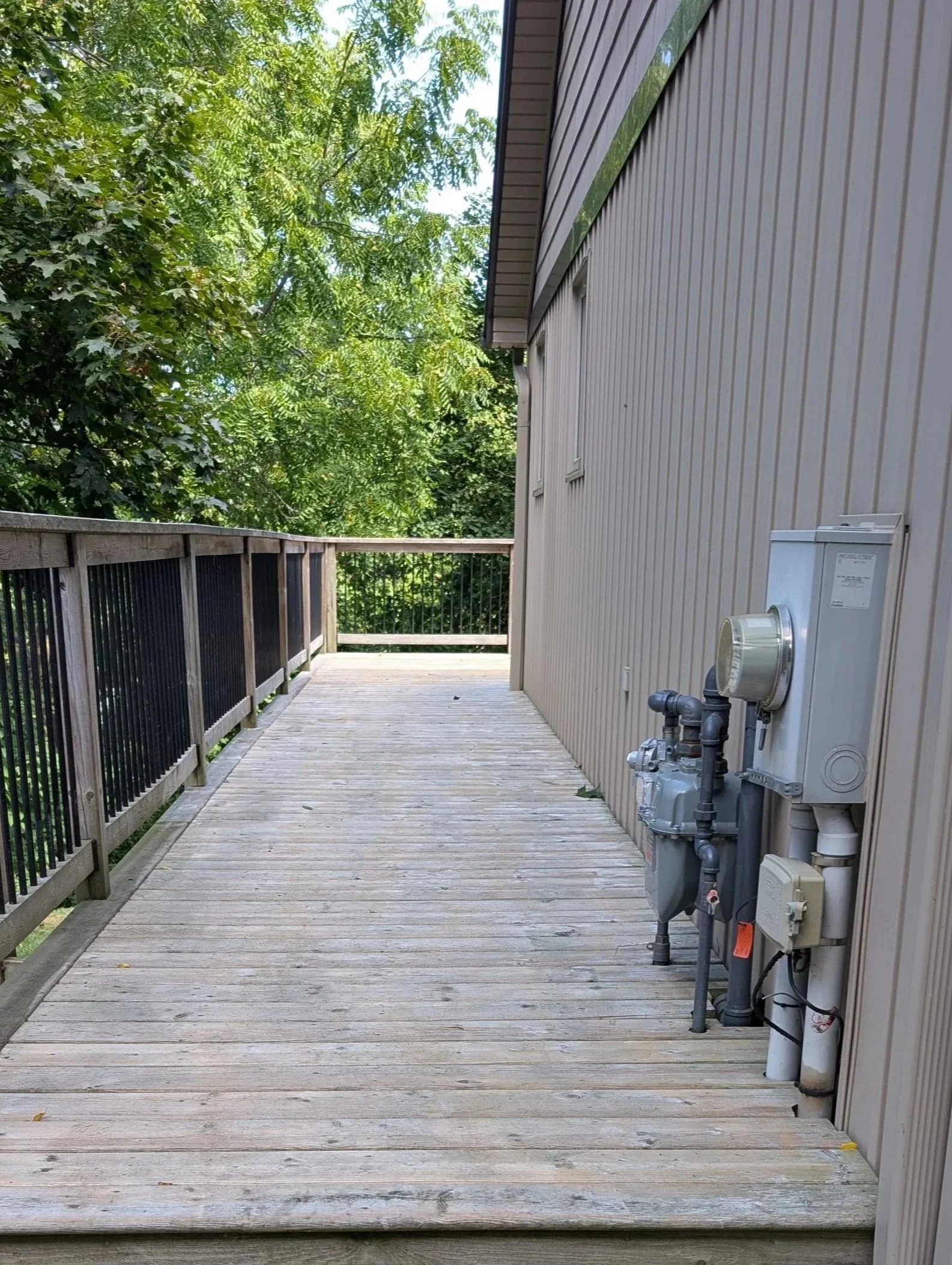 Empty wooden balcony with electrical meters and black railing, surrounded by green trees.