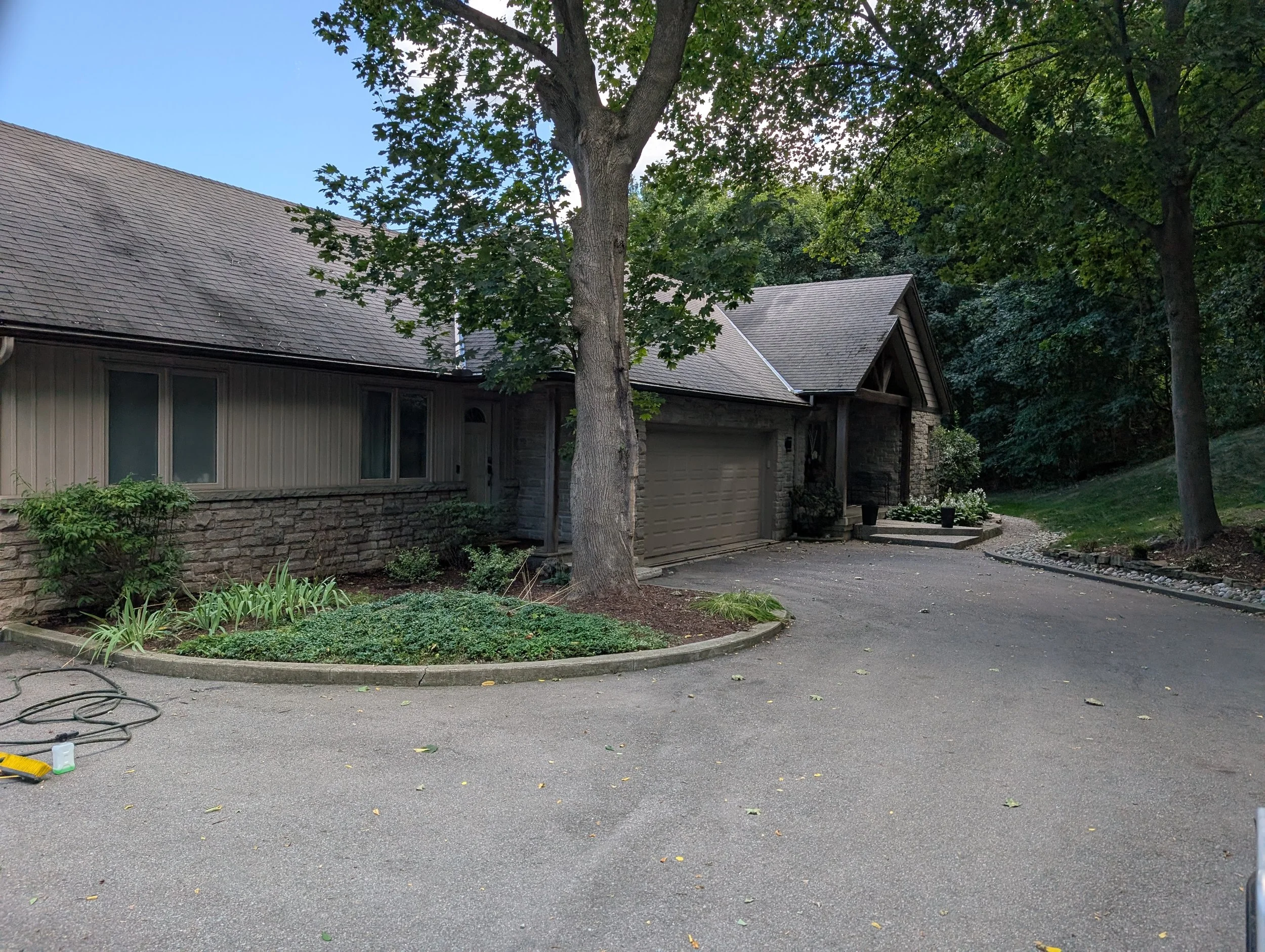 Exterior of a house with a garage, driveway, trees, and garden plants in a suburban setting during daytime.