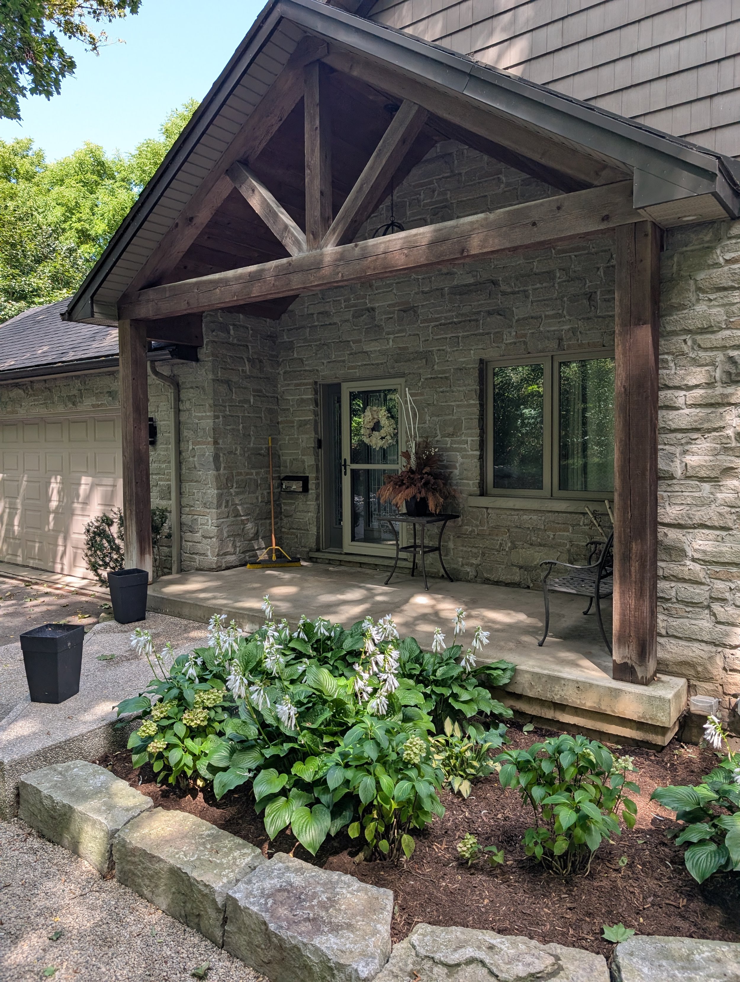 Front porch area of a house with stone walls, a concrete floor, and a wooden pergola frame. There are two black flower pots, a bench, a small round table with a floral arrangement, a window, a door, and a garden bed with white flowers and green foliage bordered by large stones.