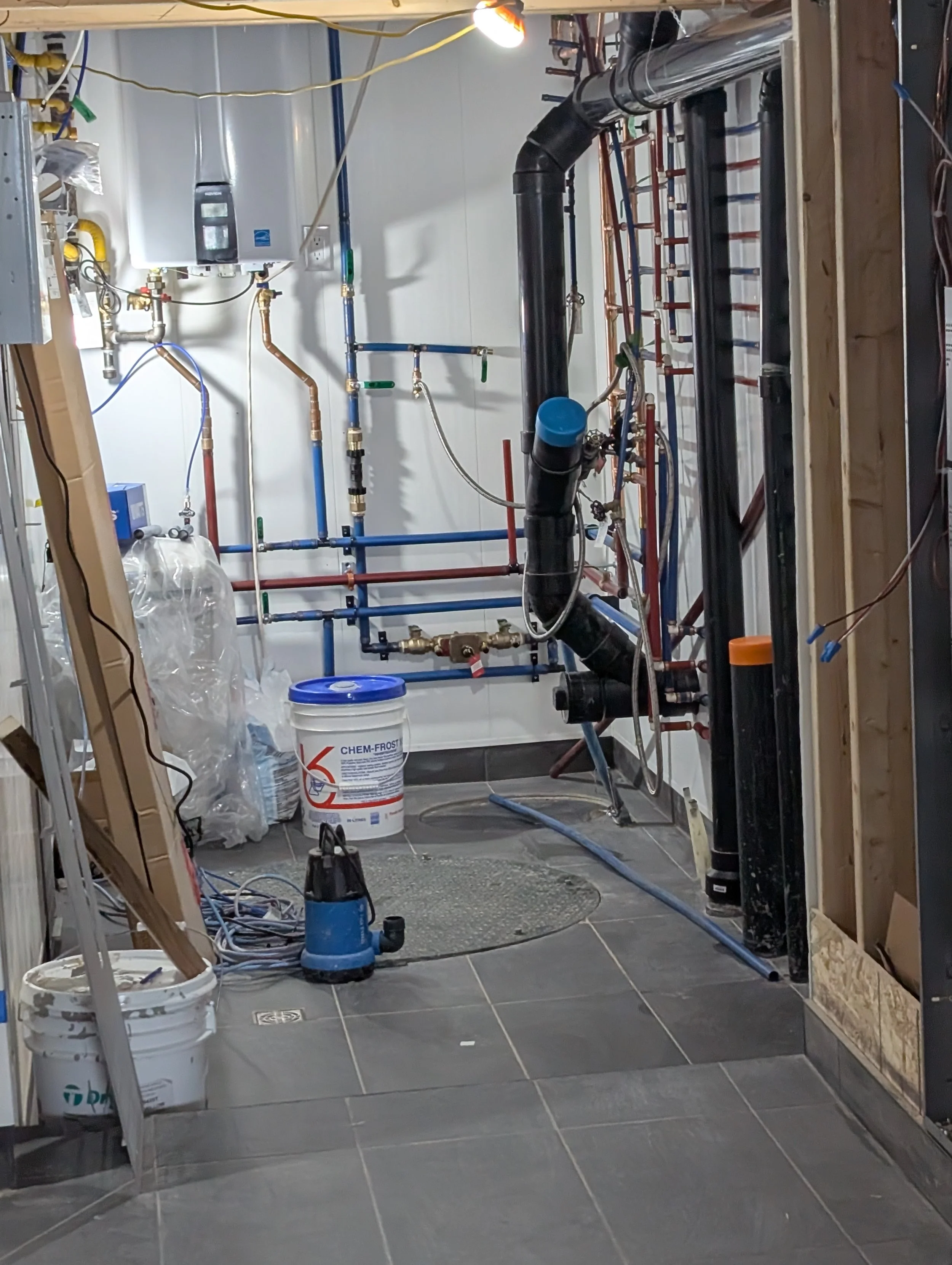 Construction site showing plumbing pipes, water heater, and construction tools in a room with tiled flooring.