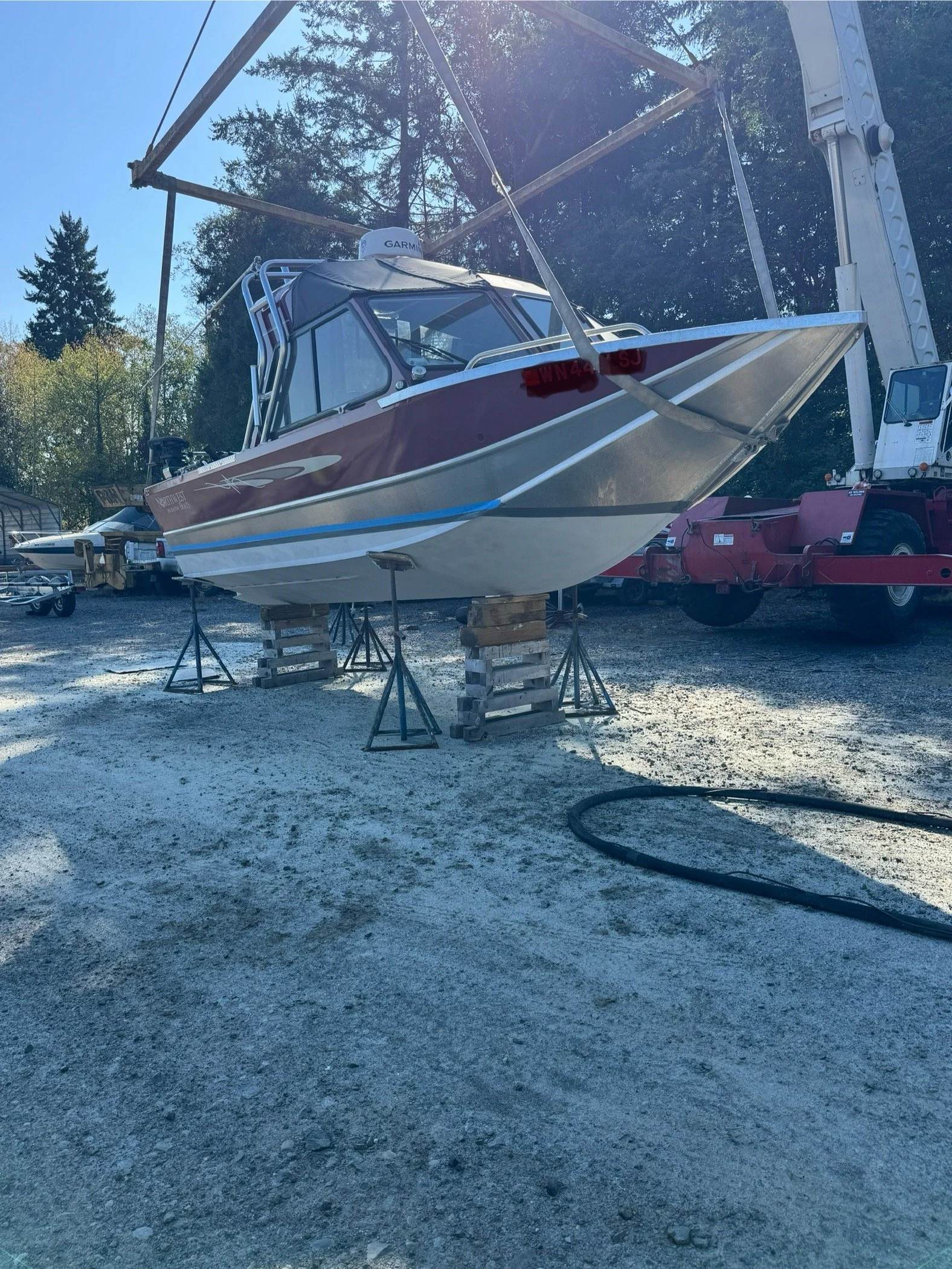 Sand Blasted A boat, it is raised on wooden supports with a crane nearby, in a boatyard surrounded by other boats and trees.