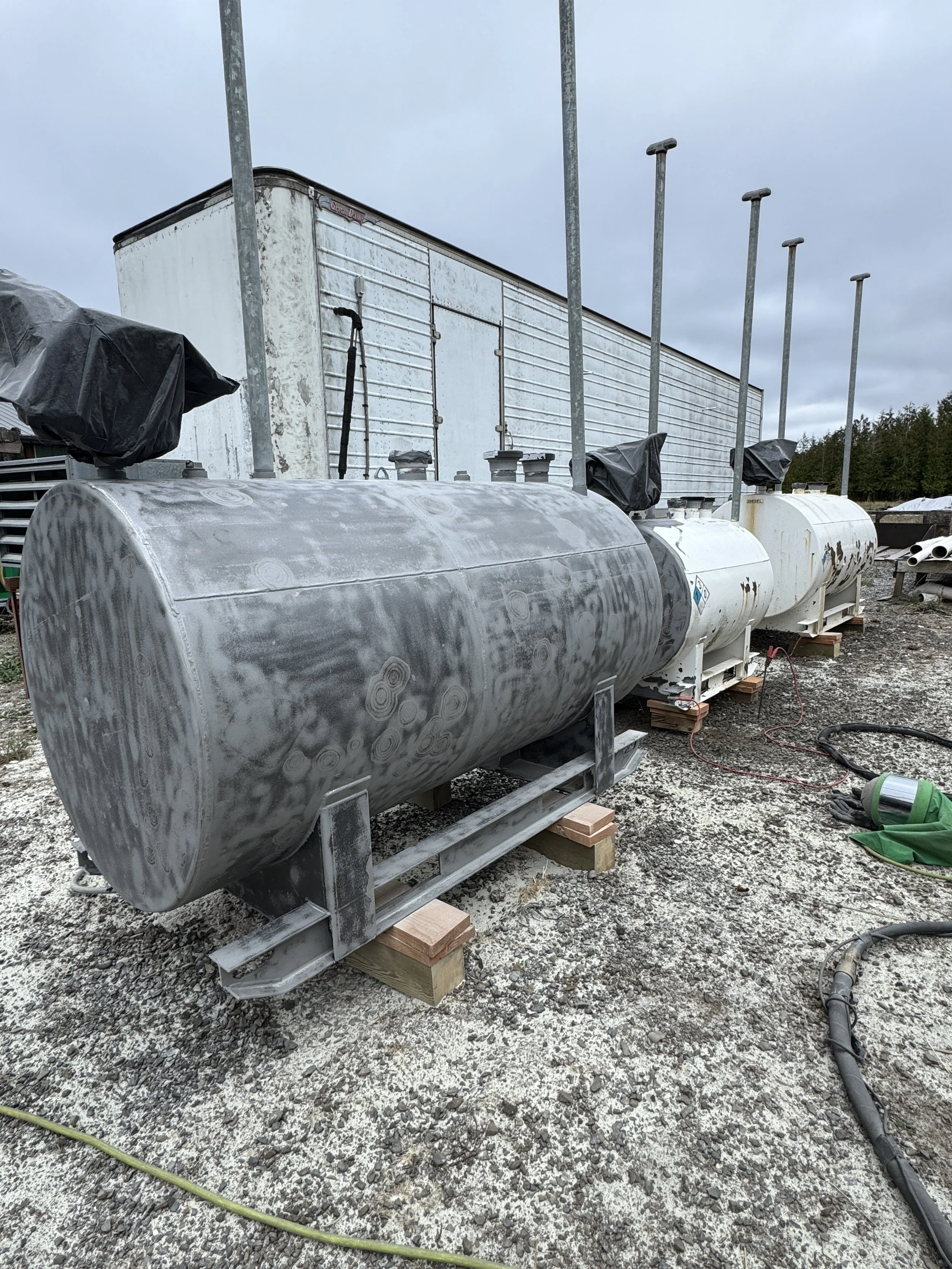 Sand blasted Four large metal storage tanks on wooden blocks at an outdoor industrial site, with a large white truck or trailer in the background, under a cloudy sky.