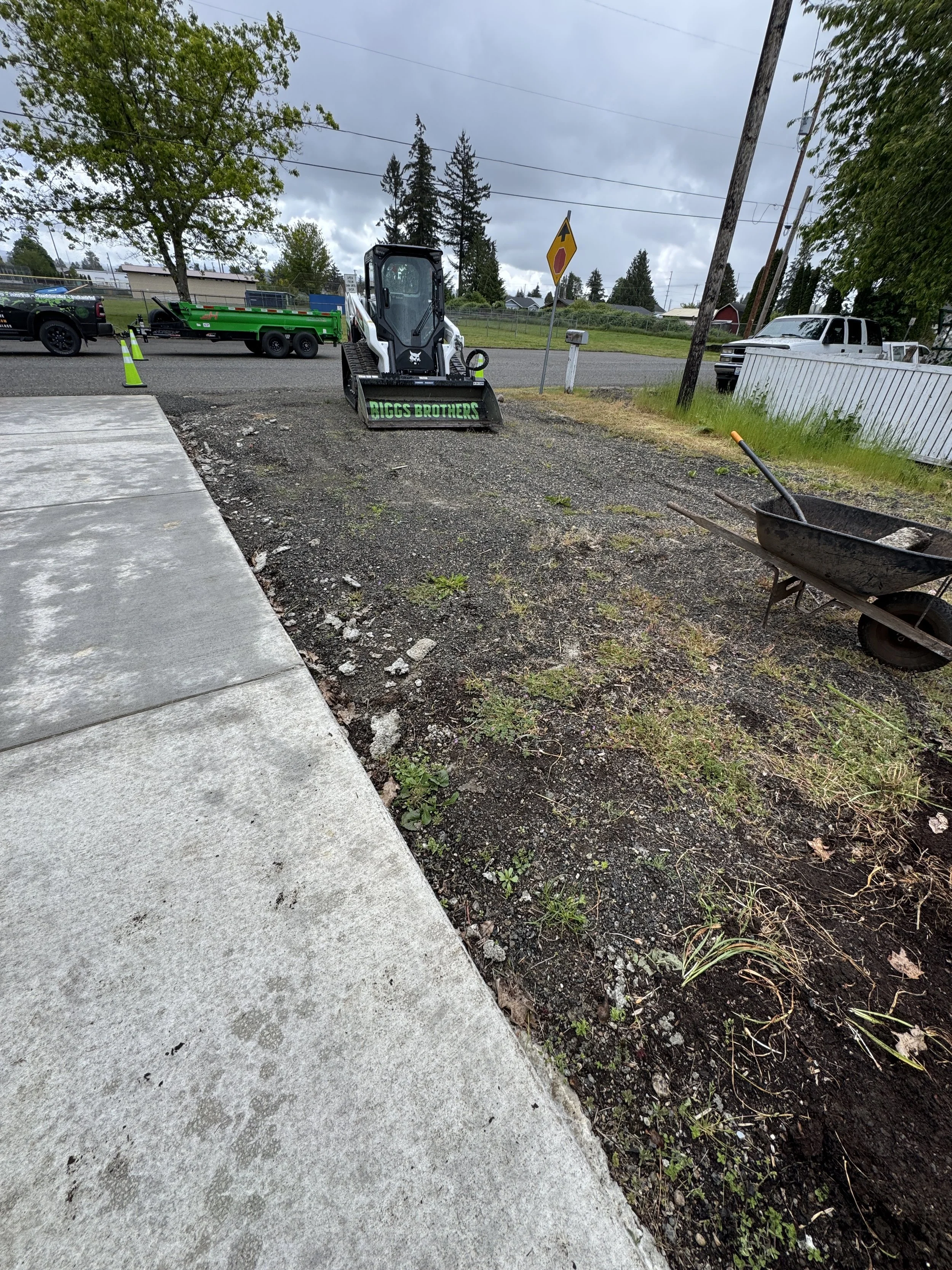 a driveway gravel job next to a yard with some grass and dirt. A small construction bulldozer with 'Biggs Brothers' written on the blade is parked at the edge of the yard. A wheelbarrow with a long handle rests nearby. 
