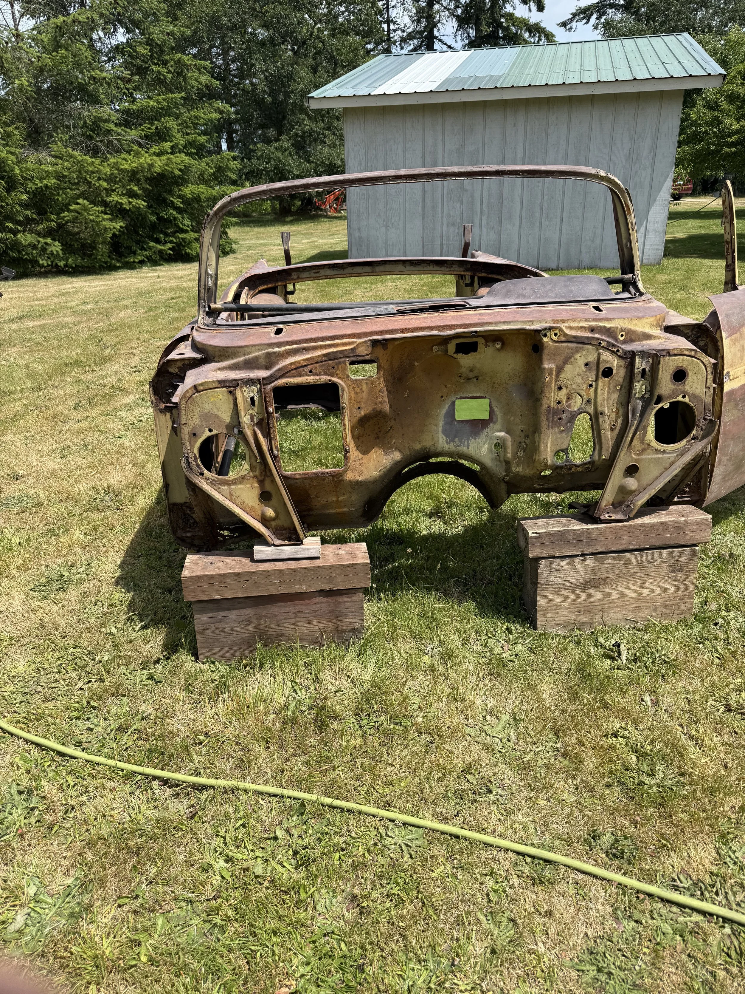 sand blasted a 57 chevy, Rusty car dashboard frame mounted on two wooden blocks outdoors, with a grassy yard, trees, and a light blue shed in the background.