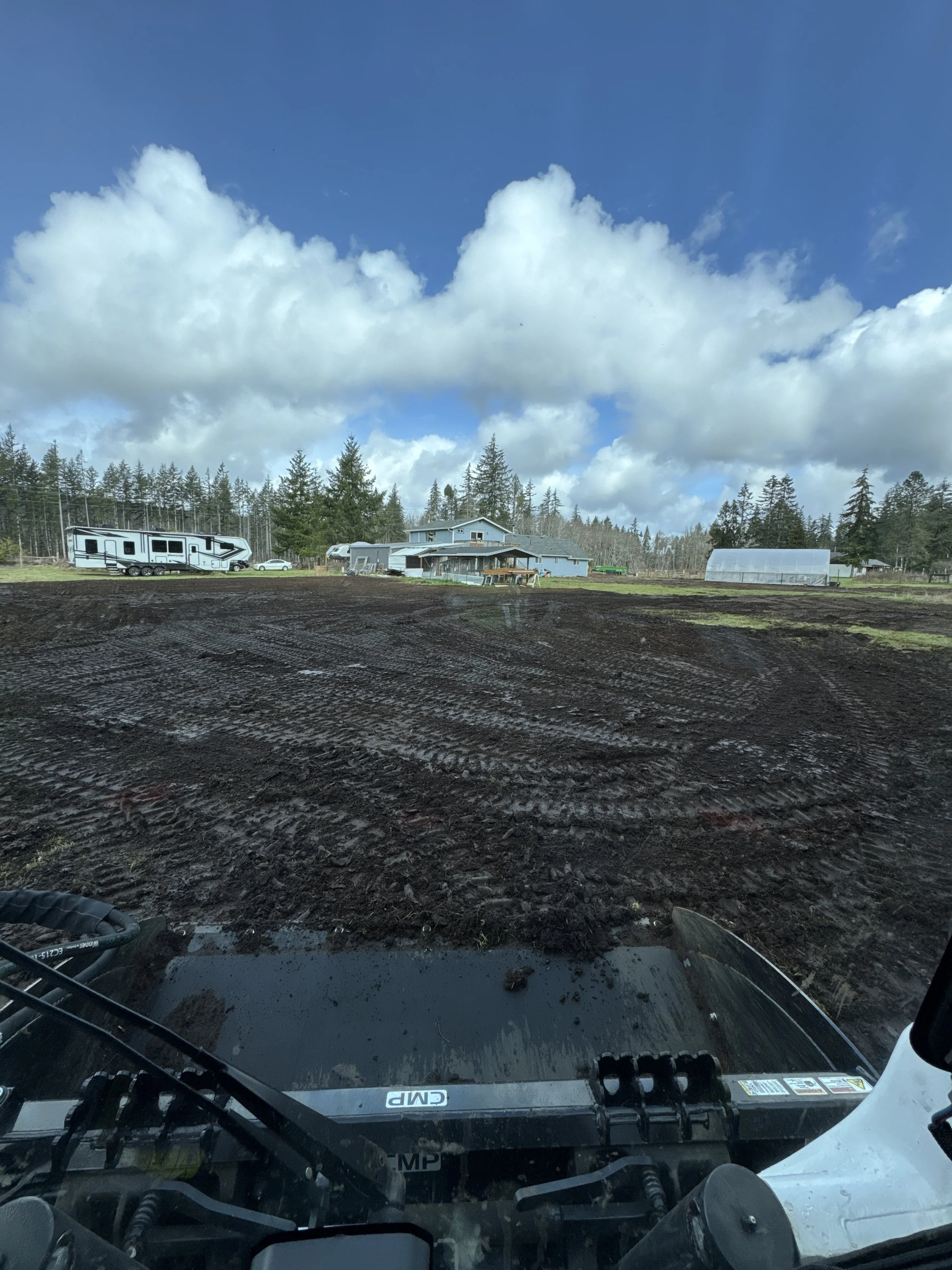 property cleared for grass, View through the windshield of a construction vehicle showing a rural farm with dark soil, greenhouses, a few buildings, a forest in the background, and a partly cloudy sky.