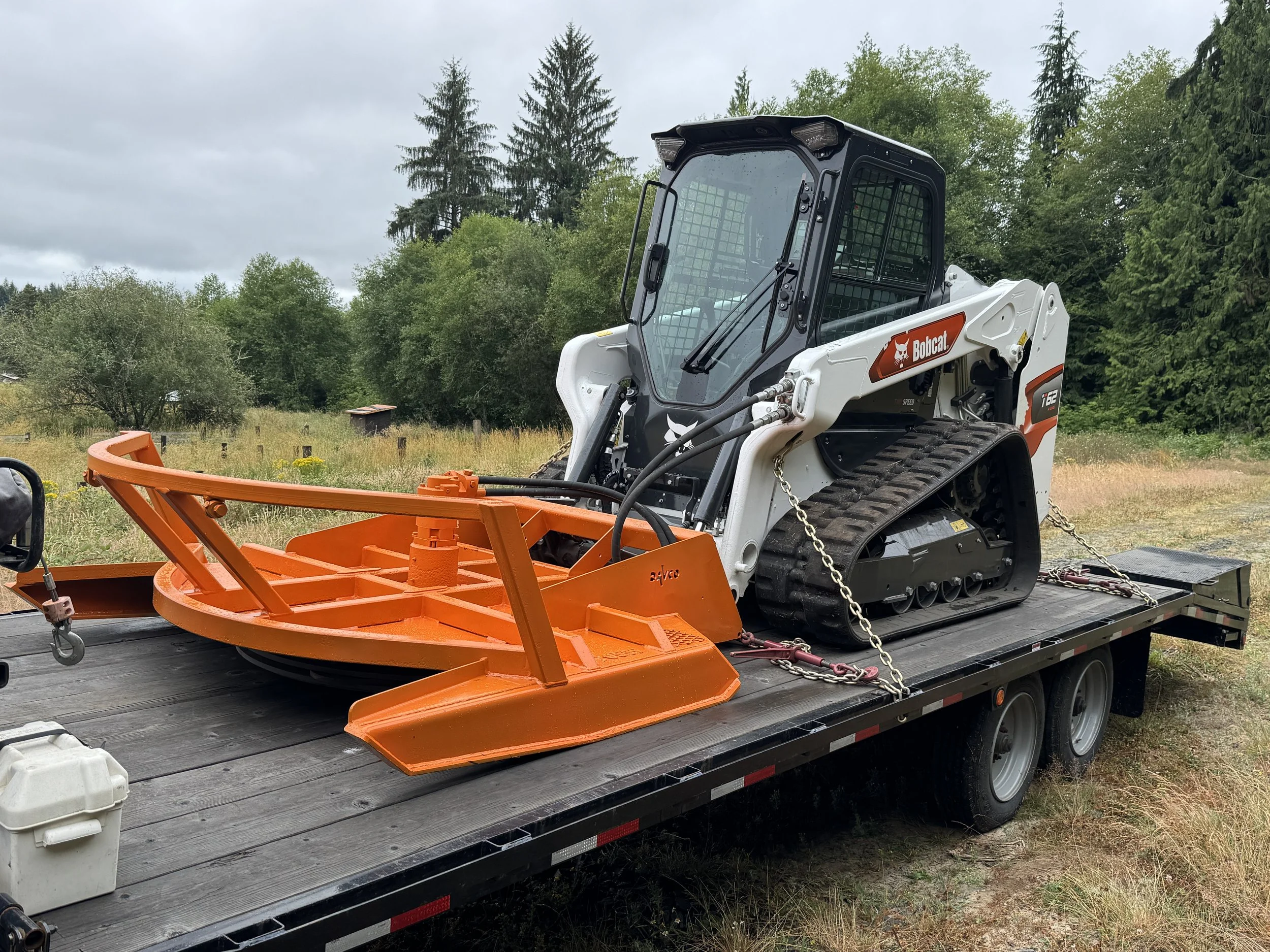 A Bobcat t62 track loader  with a Davco 72" brush hog on a flatbed trailer in an outdoor setting with grass, trees, and cloudy sky.