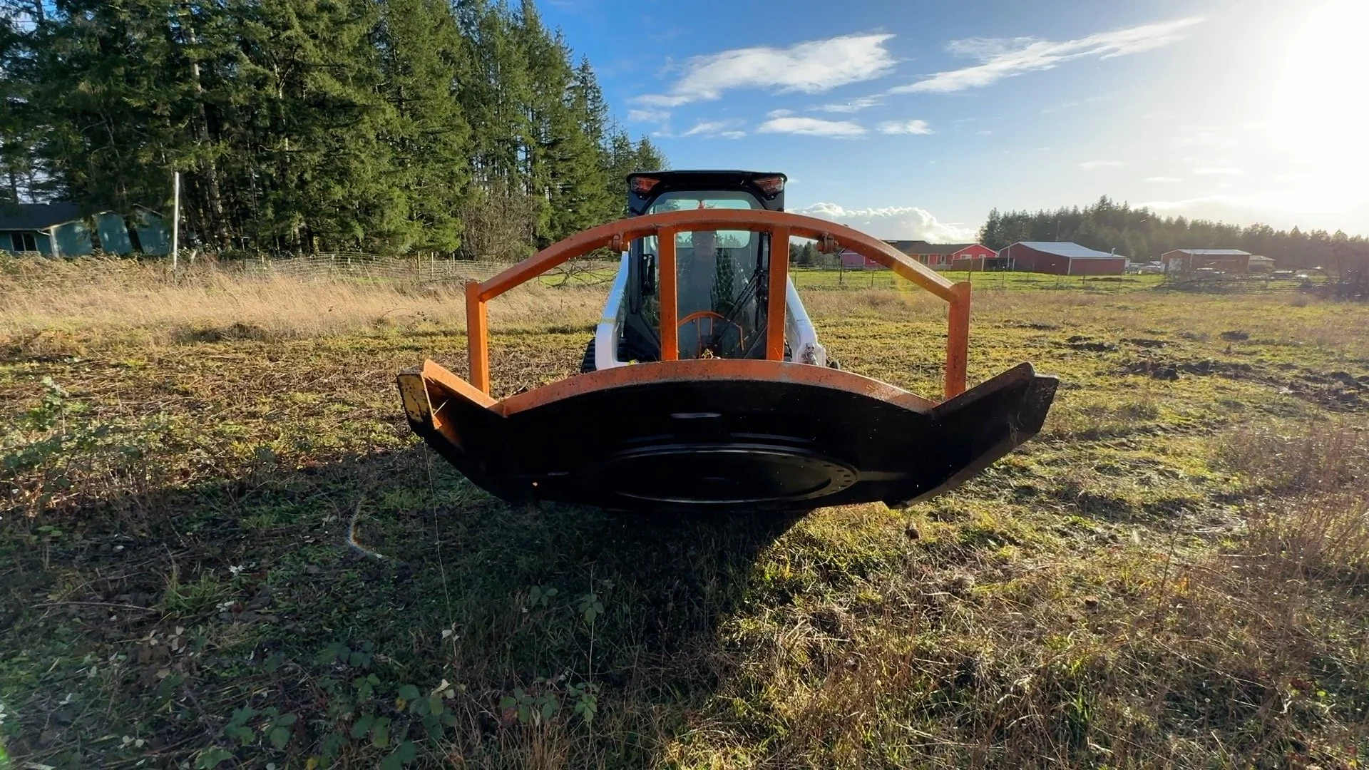 A Bobcat t62 track loader  with a Davco 72" brush hog on a grassy field, with trees and farm buildings in the distance under a partly cloudy sky.