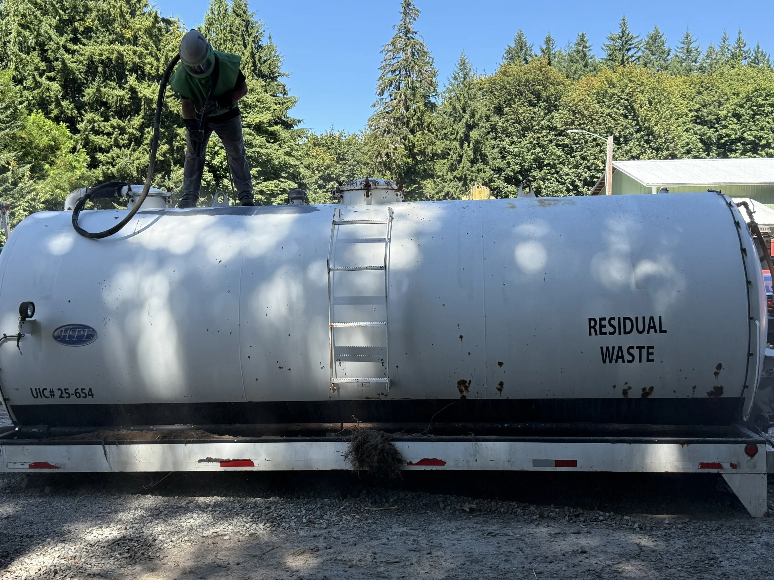 sand blasting a large white tank labeled 'Residual Waste' and 'UIC# 25-654,'. The tank is on a trailer outdoors, surrounded by trees and clear blue skies.