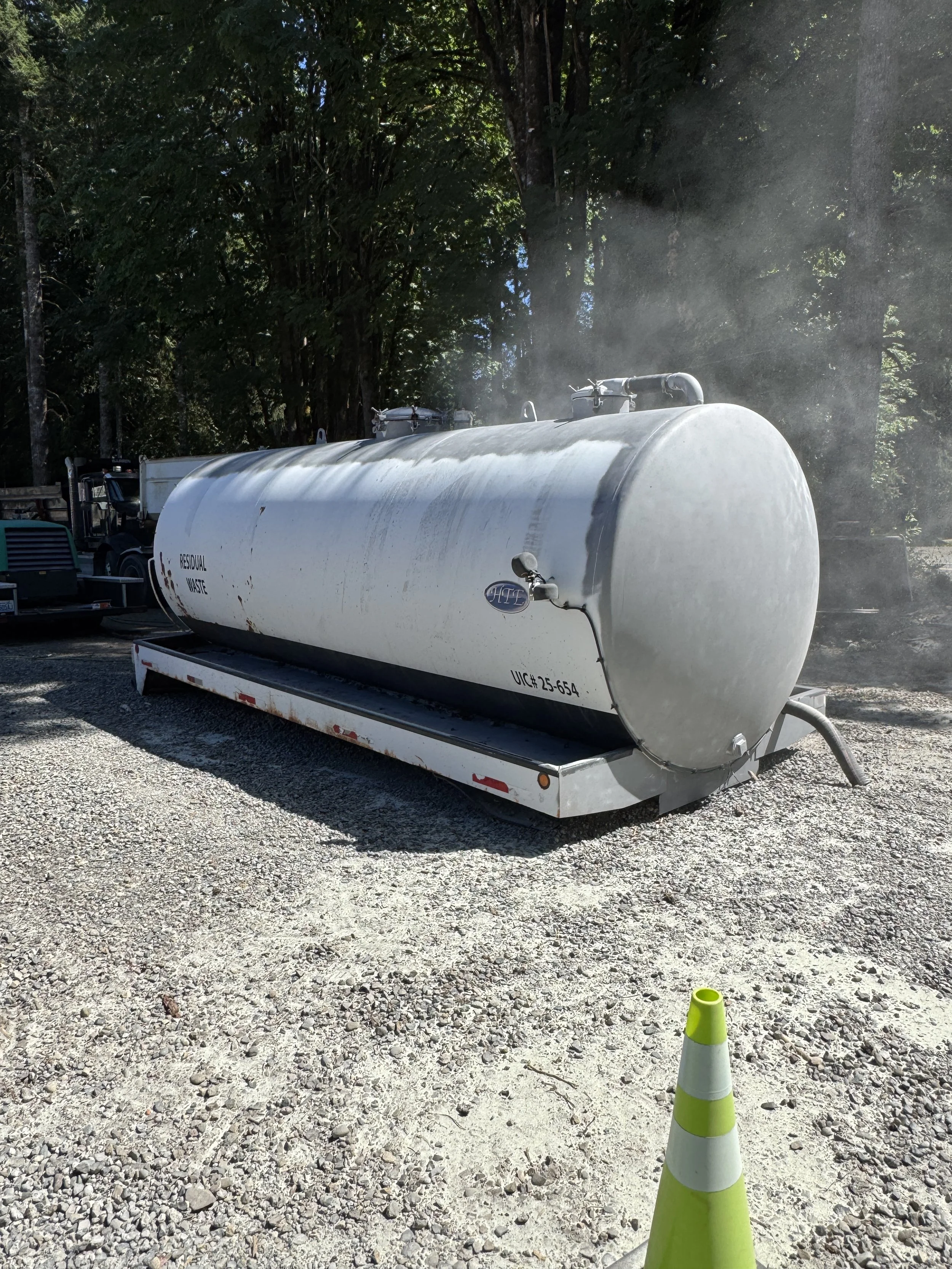 Large white sand blasted residual waste tank mounted on a trailer, emitting steam or vapor, with green trees in the background and a bright traffic cone in the foreground.
