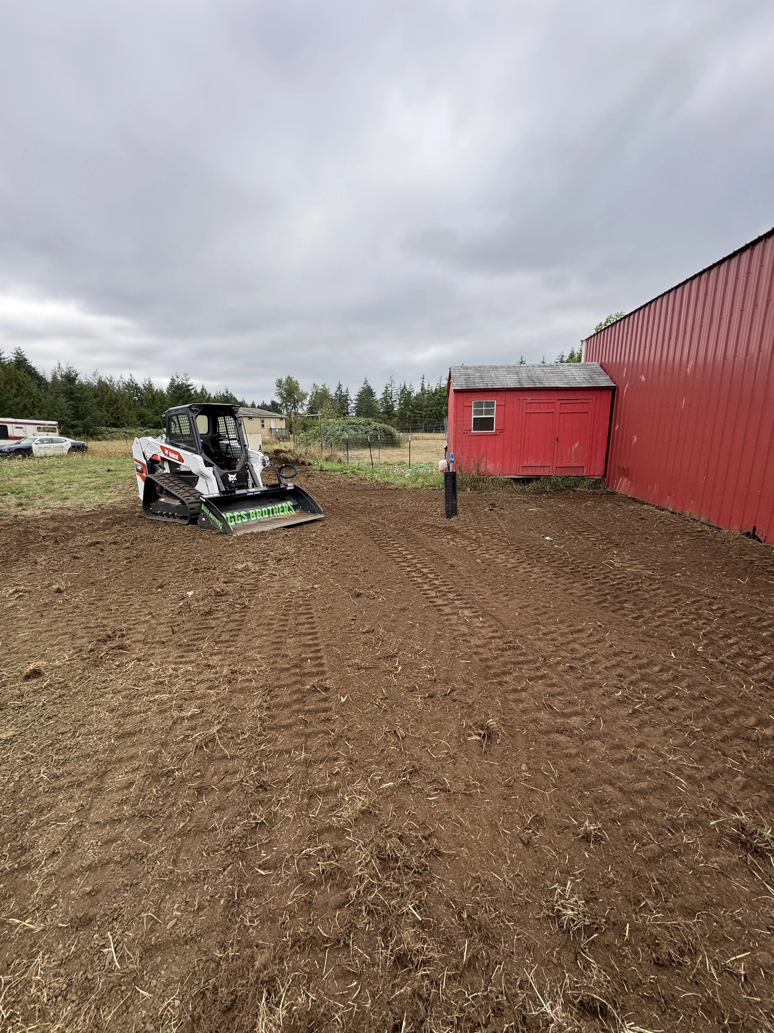 A A Bobcat t62 track loader  with a Davco 72" brush hog parked on freshly stripped dirt area near a red building on a cloudy day.