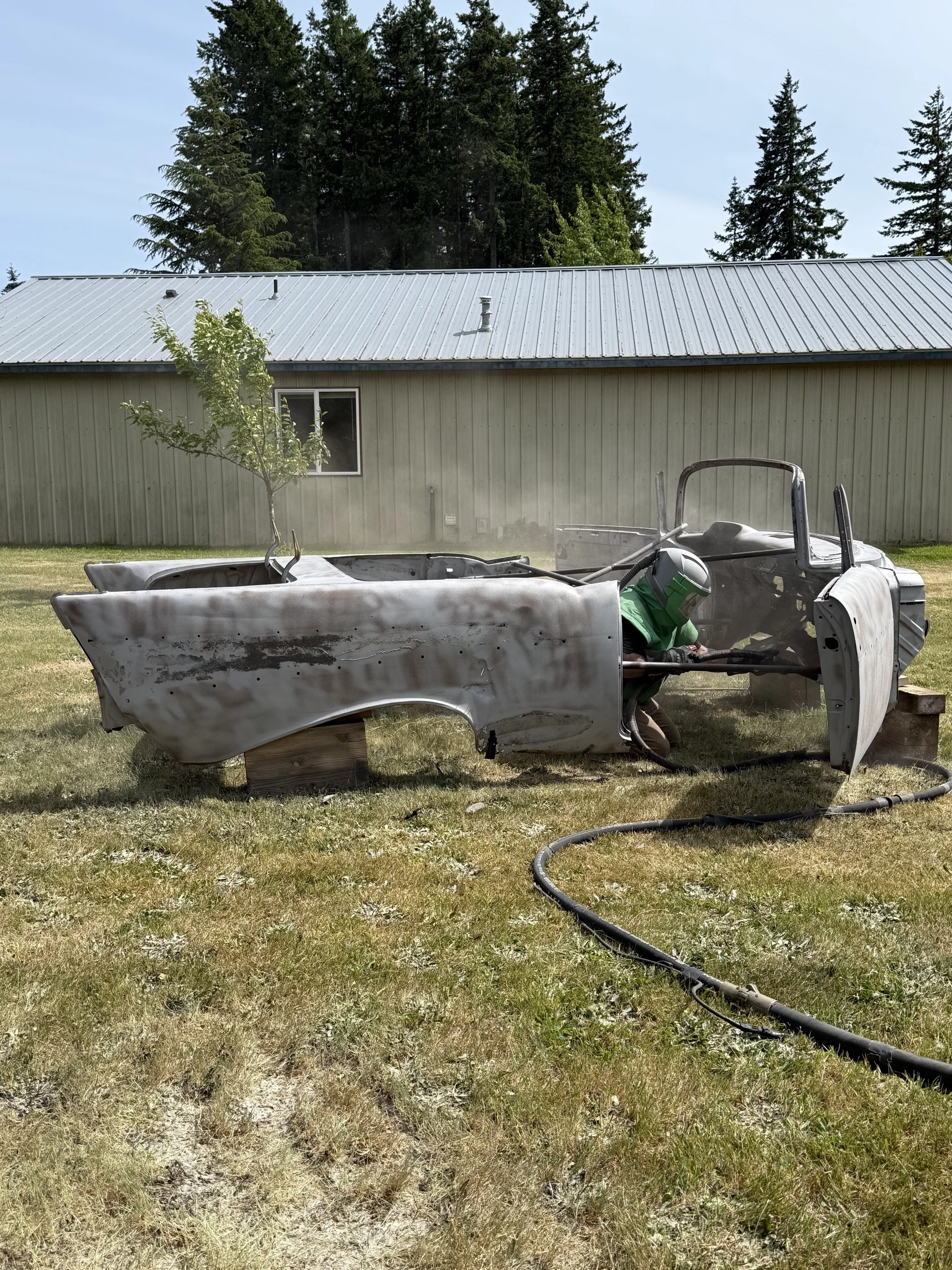 sand blasted a 57 chevy, Person wearing green protective gear, including a welding helmet and gloves, working on a rusted, damaged vintage car body with a tree growing inside it on the grass near a metal building and surrounded by trees.
